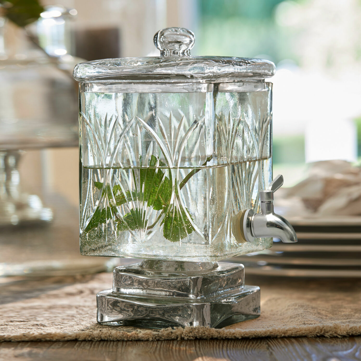 Glazen drankendispenser met deksel en kraan, versierd met patroon en groene bladeren.