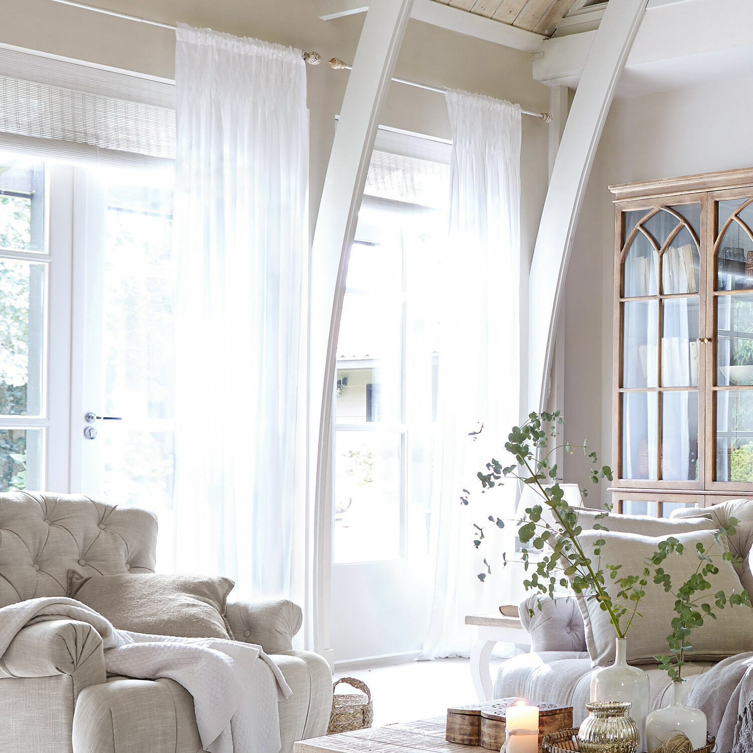 Gordijn Otilie Lichte woonkamer met beige gestoffeerde stoel, witte gordijnen en vintage houten vitrinekast
