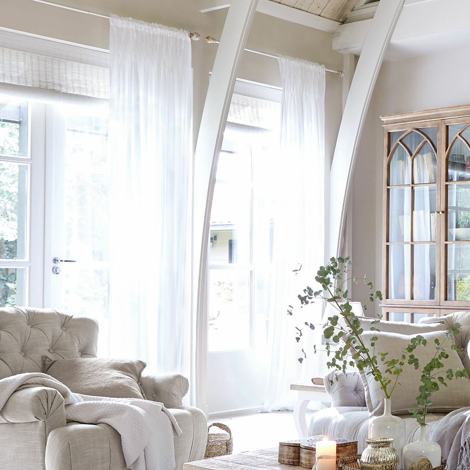 Gordijn Otilie Lichte woonkamer met beige gestoffeerde stoel, witte gordijnen en vintage houten vitrinekast