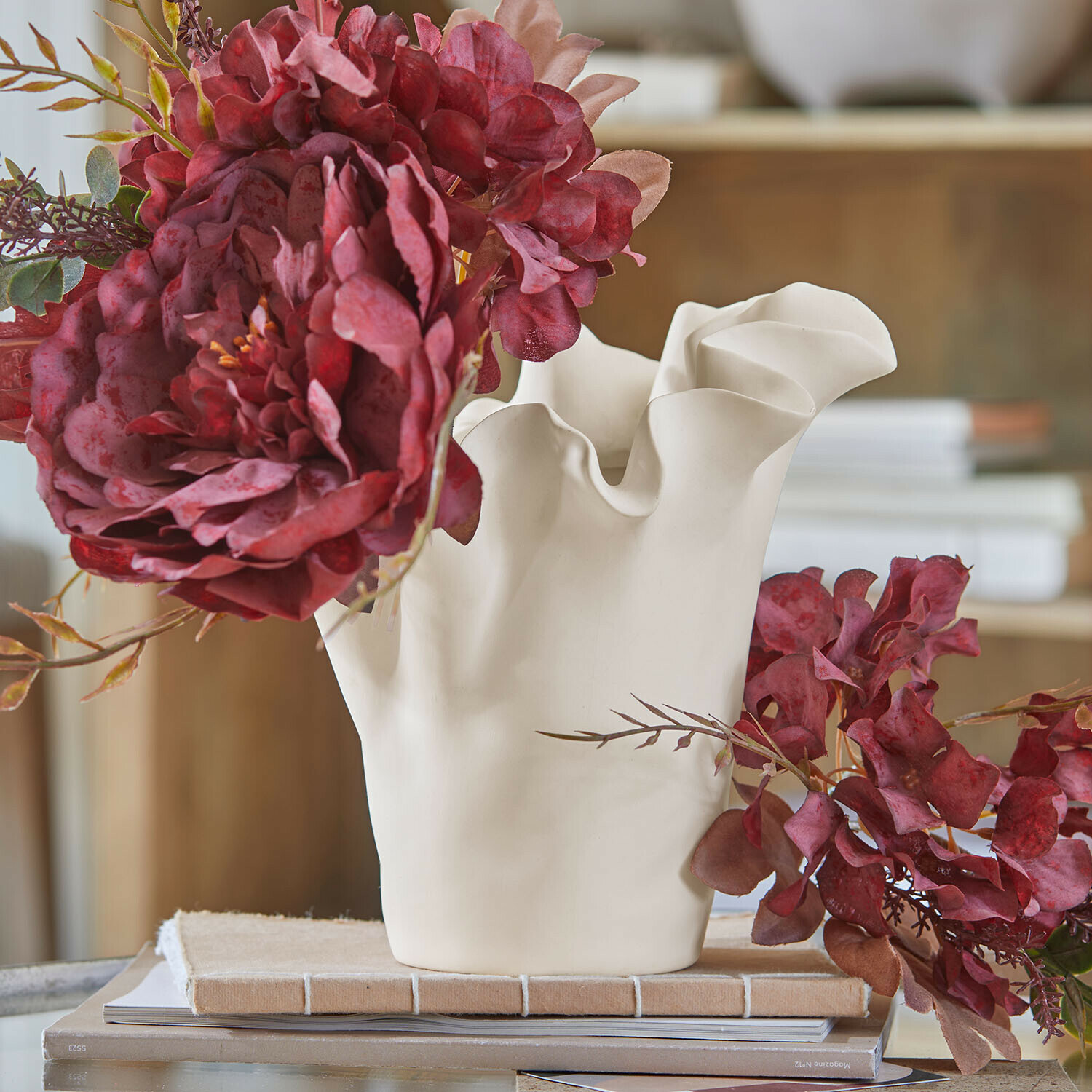 Witte keramische vaas met golvende rand en rode kunstbloemen op tafel met boeken