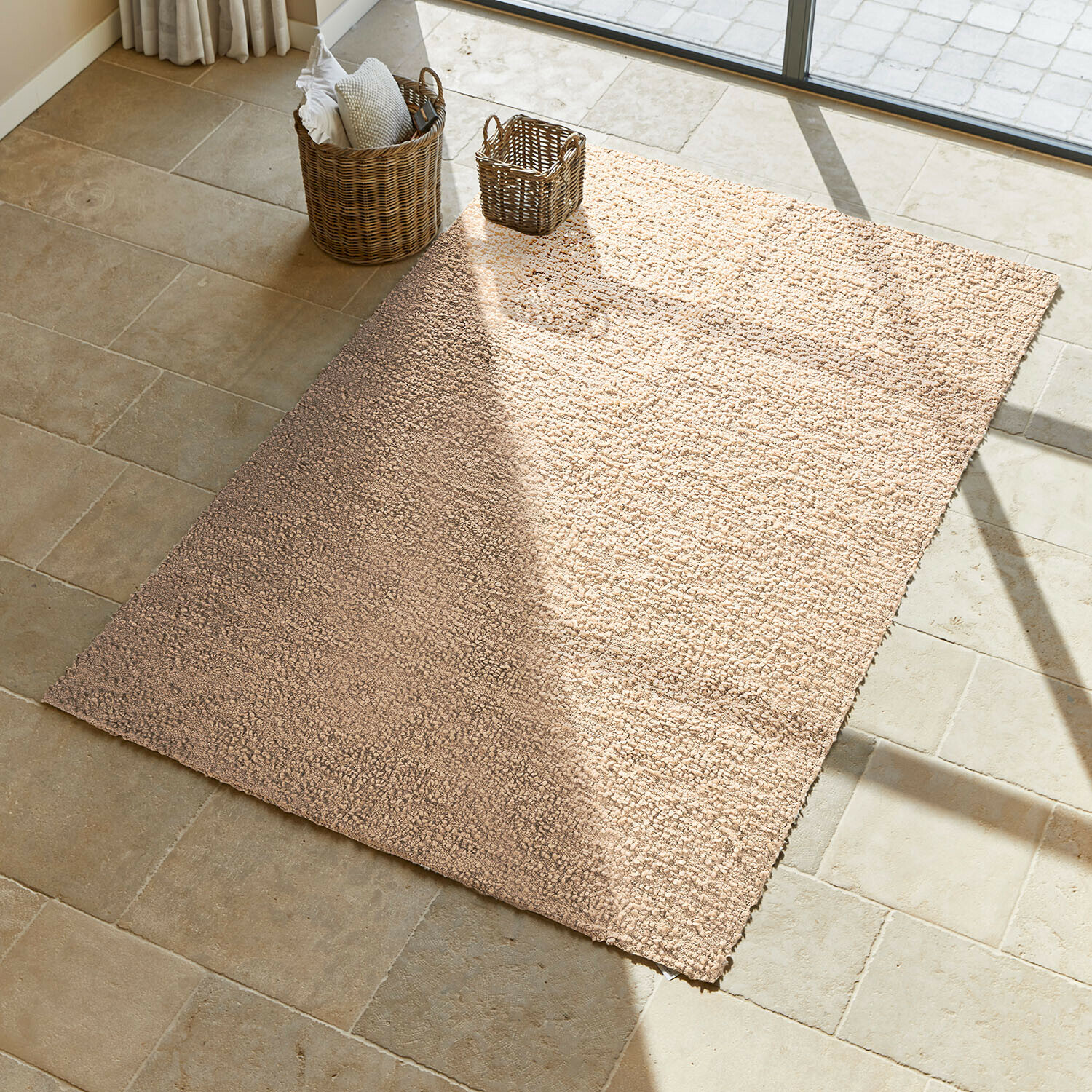 Vloerkleed Wodaha Beige vloerkleed van zacht materiaal op lichte stenen vloer met rieten manden ernaast