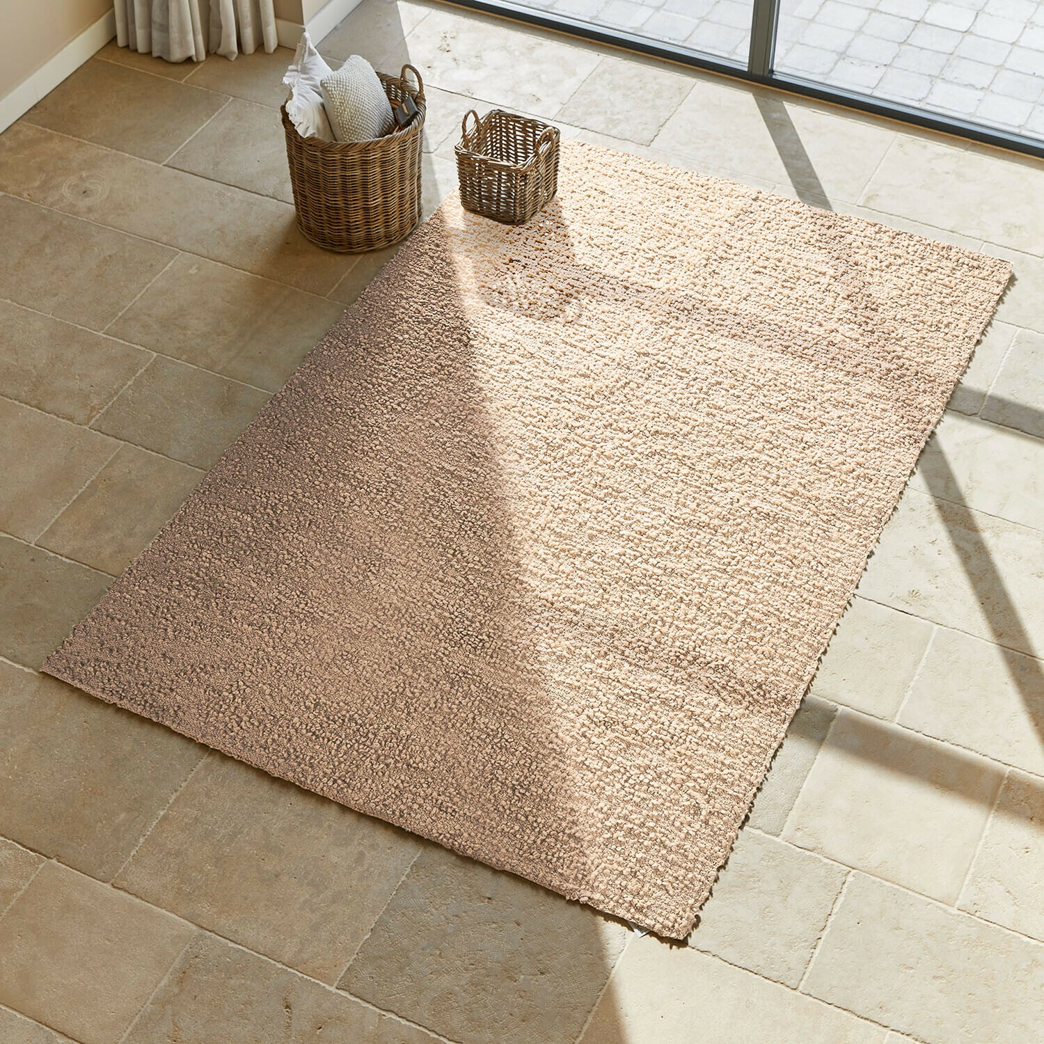 Vloerkleed Wodaha Beige vloerkleed van zacht materiaal op lichte stenen vloer met rieten manden ernaast