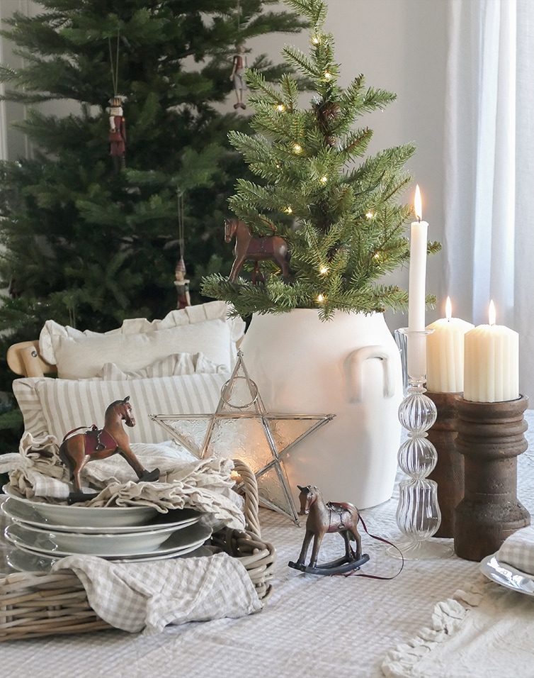 Kerstdecoratie met witte kan, groene kerstboom, kaarsen en houten hobbelpaardjes op tafel