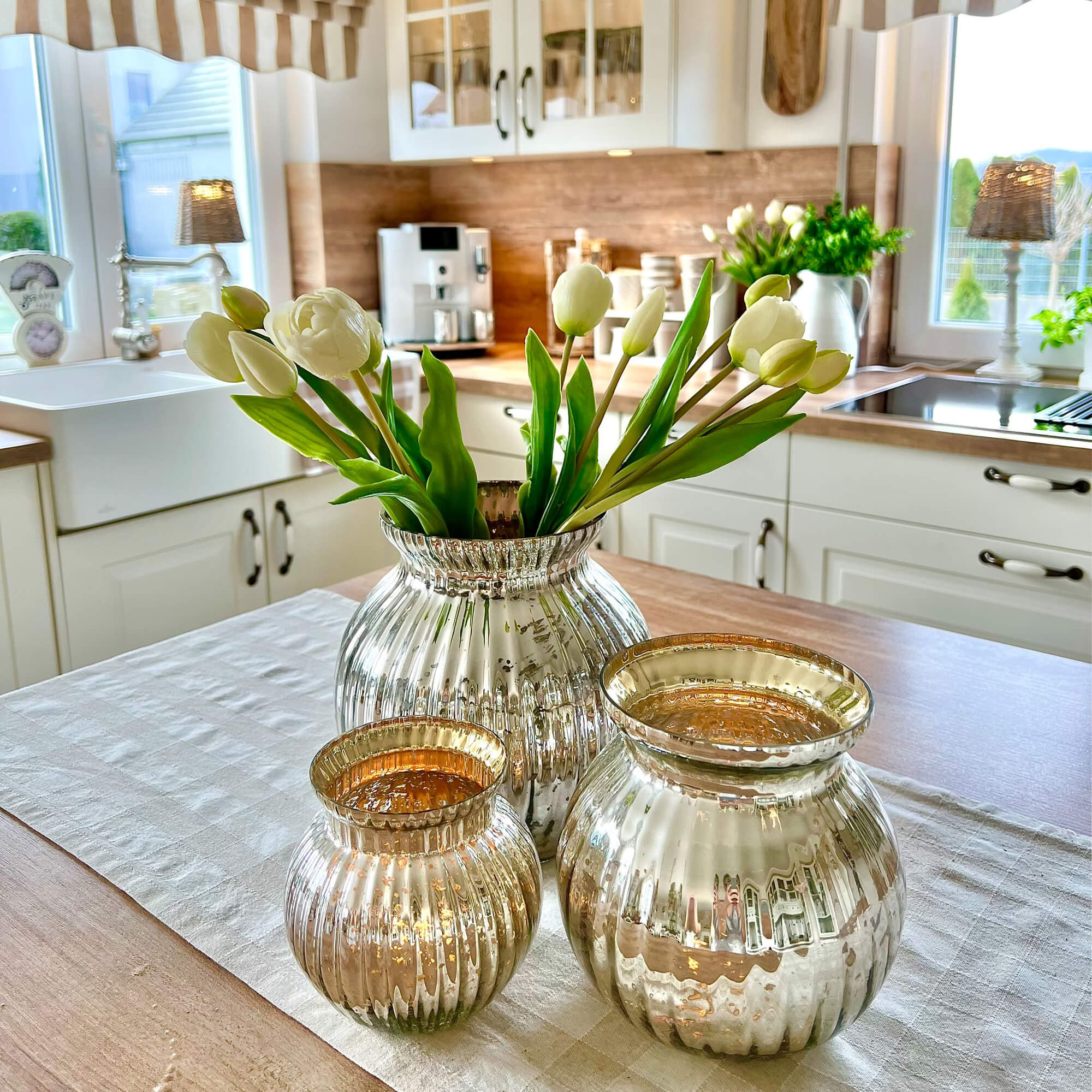 Set van drie zilverkleurige glazen vazen op houten tafel in keuken met witte tulpen