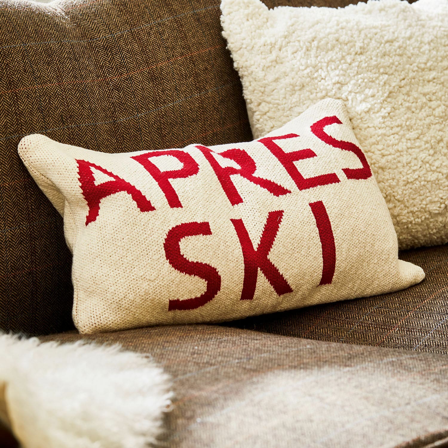 Kussenhoes Apres Ski Beige kussen met rode tekst Apres Ski op bruine bank met zachte bontdeken