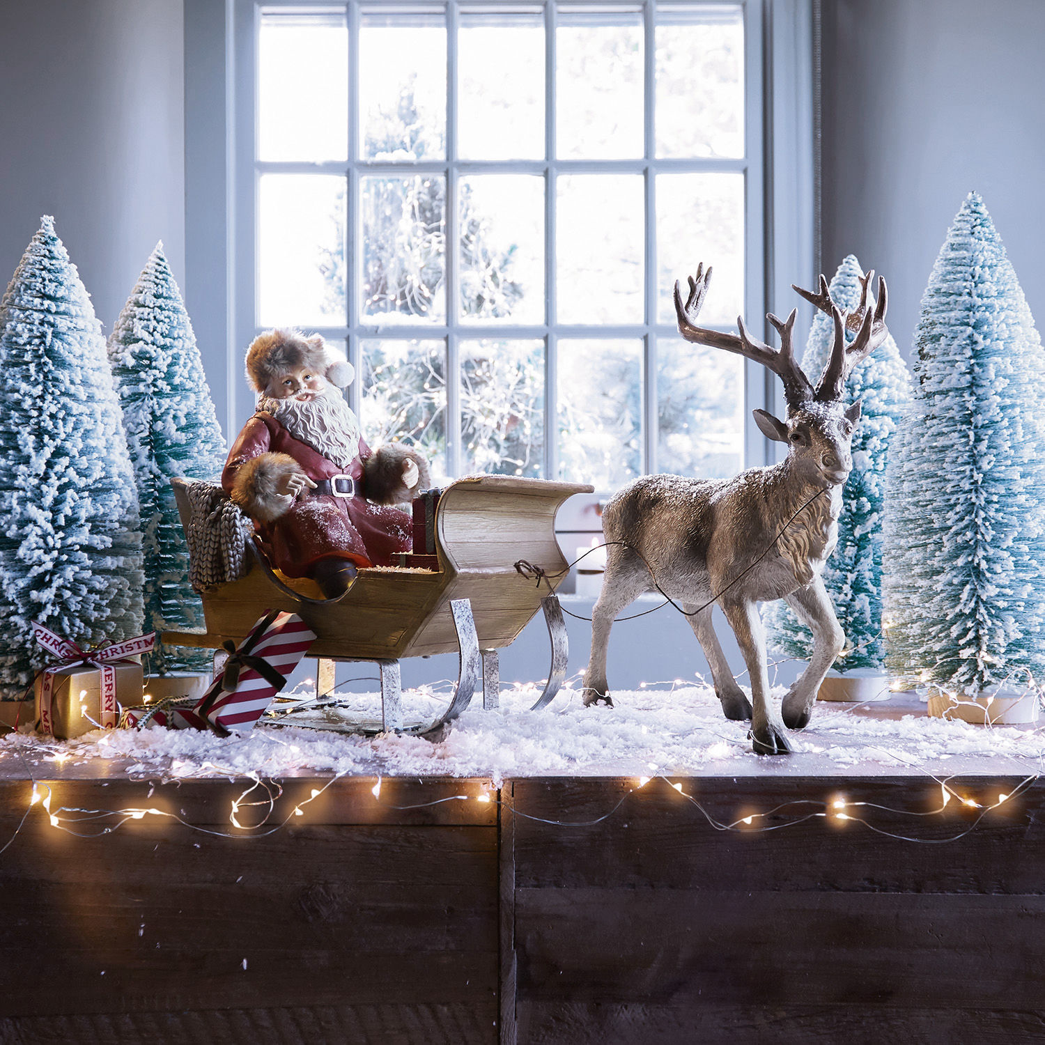Kerstmanfiguur in rode mantel op houten slee met rendier en besneeuwde dennenbomen als decoratie