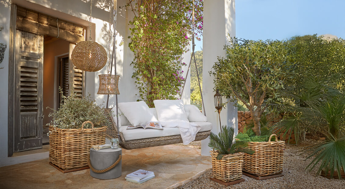 Rotan schommel met witte kussens op terras met rotan lampen en planten in gevlochten manden