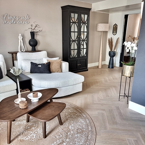 Gezellige woonkamer met lichtgrijze hoekbank, zwarte vitrinekast en houten salontafel op ronde beige vloerkleed