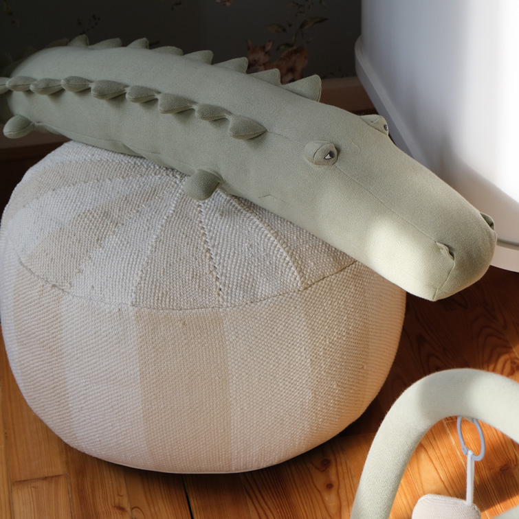 Zachte lichtgroene pluche krokodil op een beige geweven poef op een houten vloer, kinderkamerdecoratie