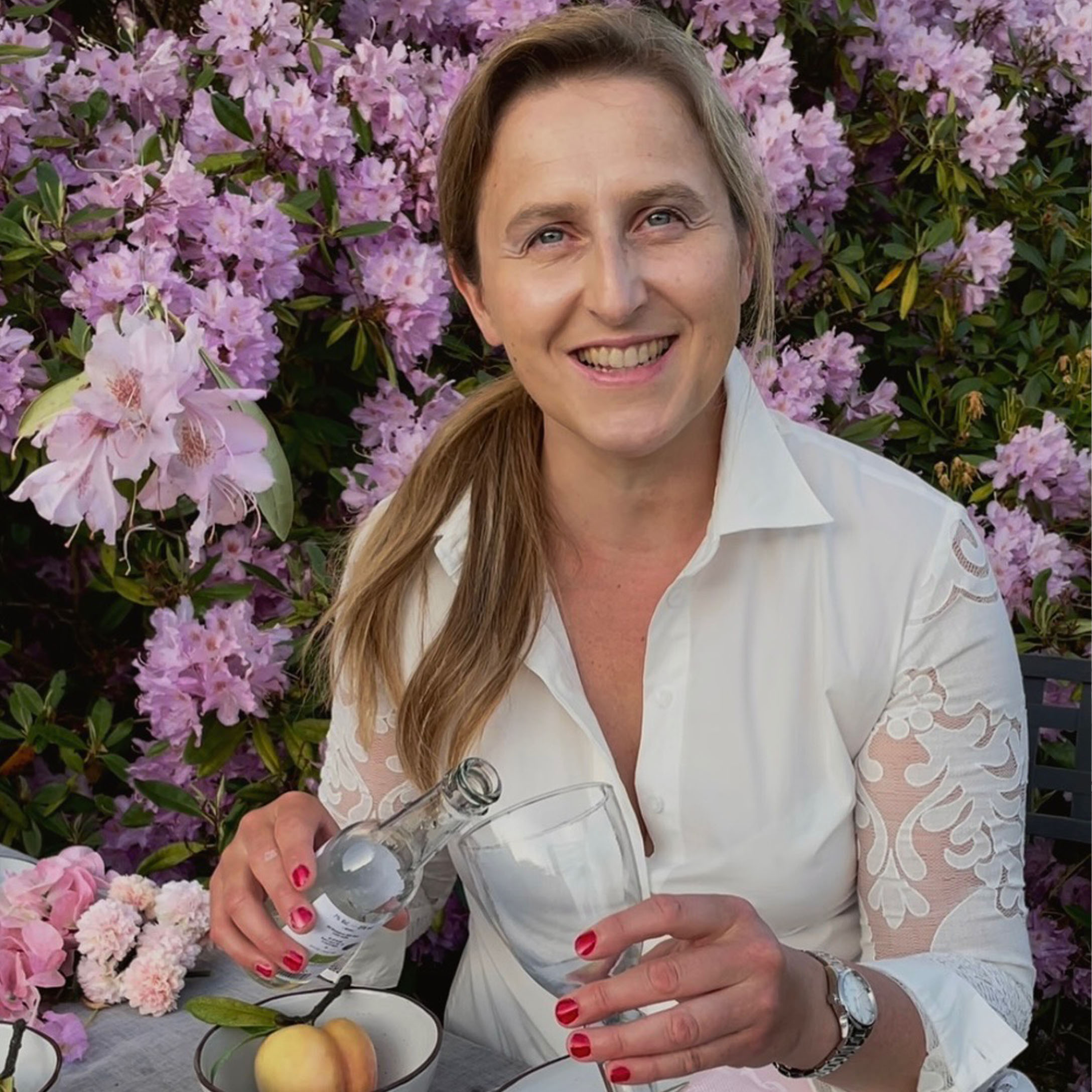 Vrouw in wit overhemd met kanten mouwen schenkt drankje in glas voor roze bloemen