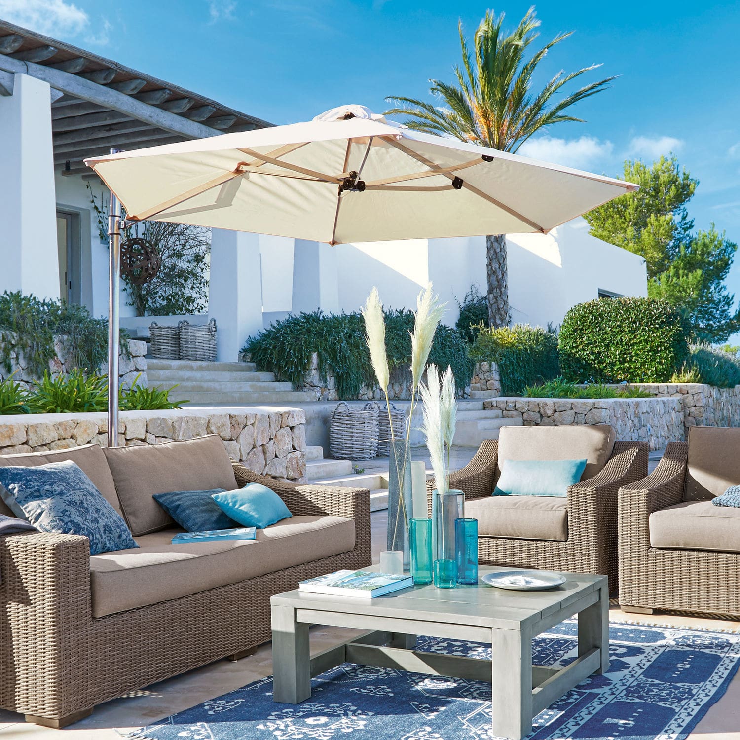Tuinmeubelset van beige kunststof rattan met crèmekleurige parasol op terras met blauw tapijt