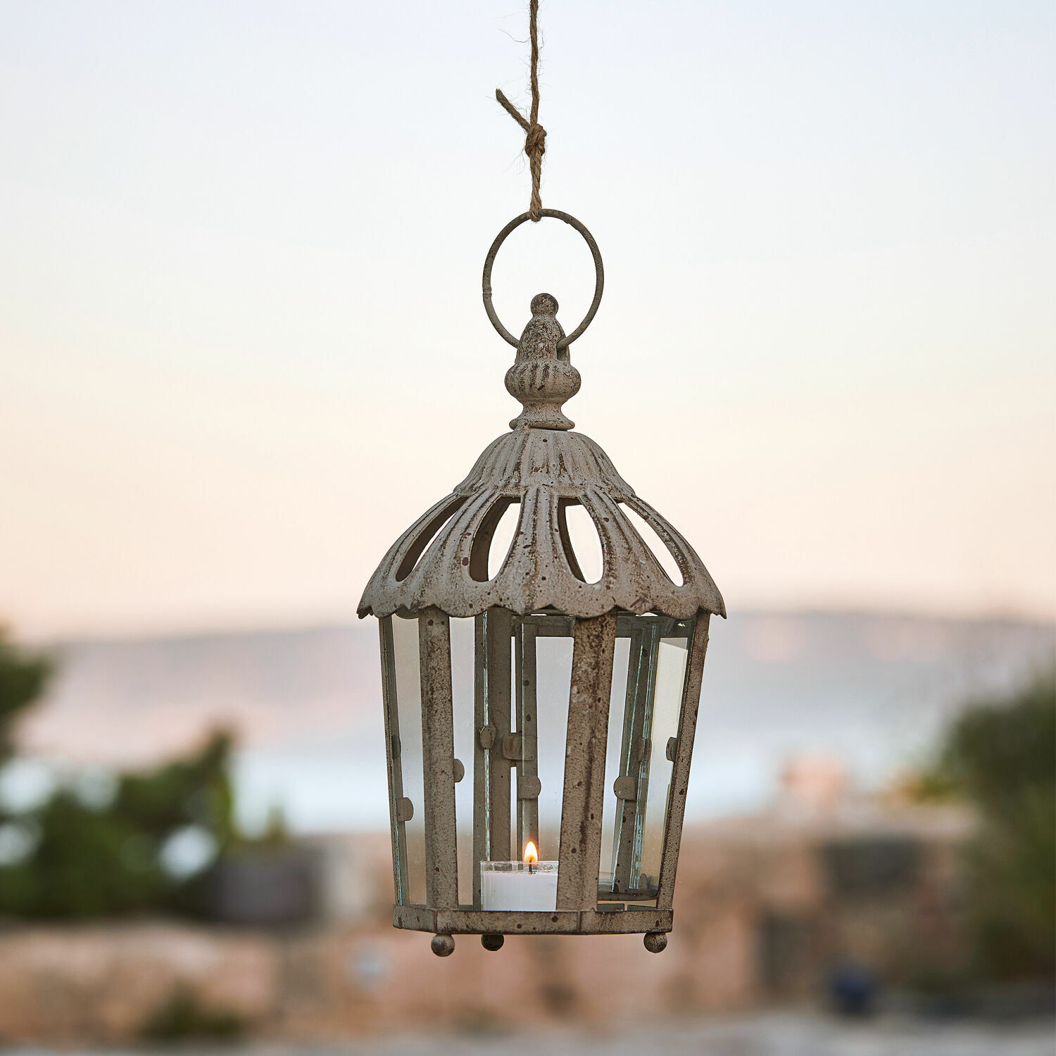 Lantaarn Artemisie Hangende metalen lantaarn in vintage stijl in verweerd beige met helder glas en brandende kaars.