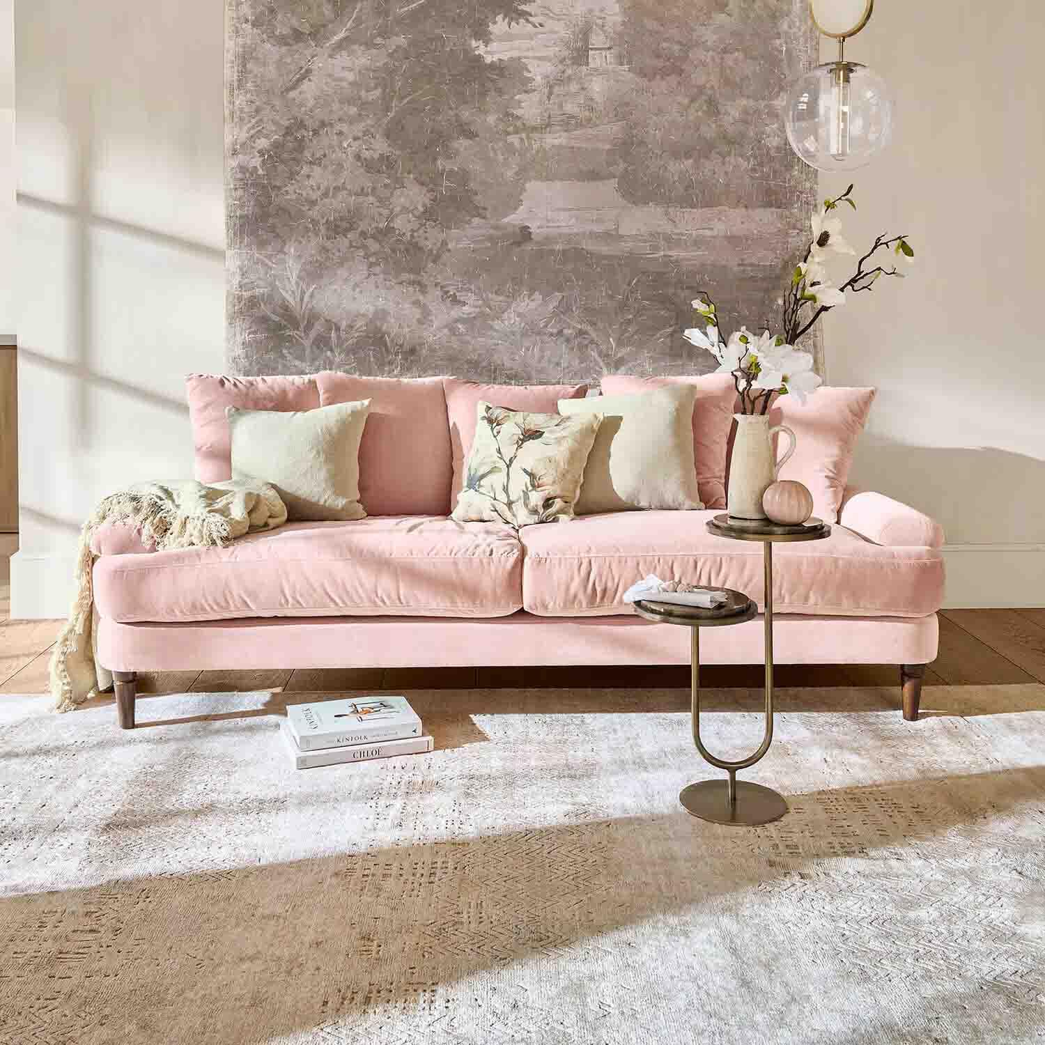 Roze fluwelen bank met kussens en bijzettafel in lichte woonkamer met wanddecoratie