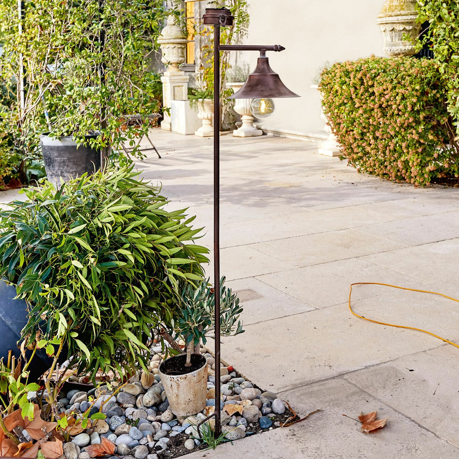 Lantaarn op zonne-energie Kaley Metalen tuinlamp in roestkleur met gloeilamp, staand op stenen tussen groene planten