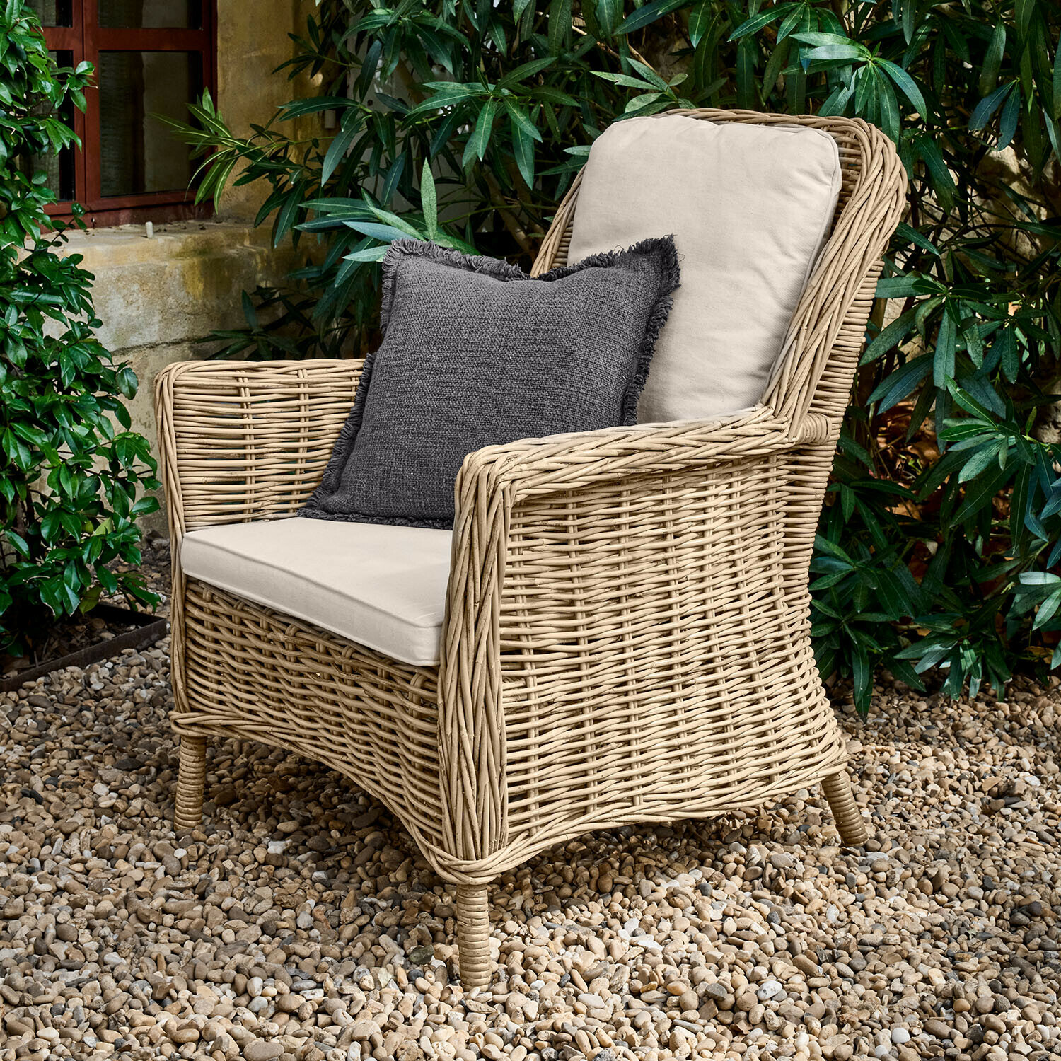 Rotan fauteuil met crèmekleurige kussens en donkergrijs sierkussen op grindterras