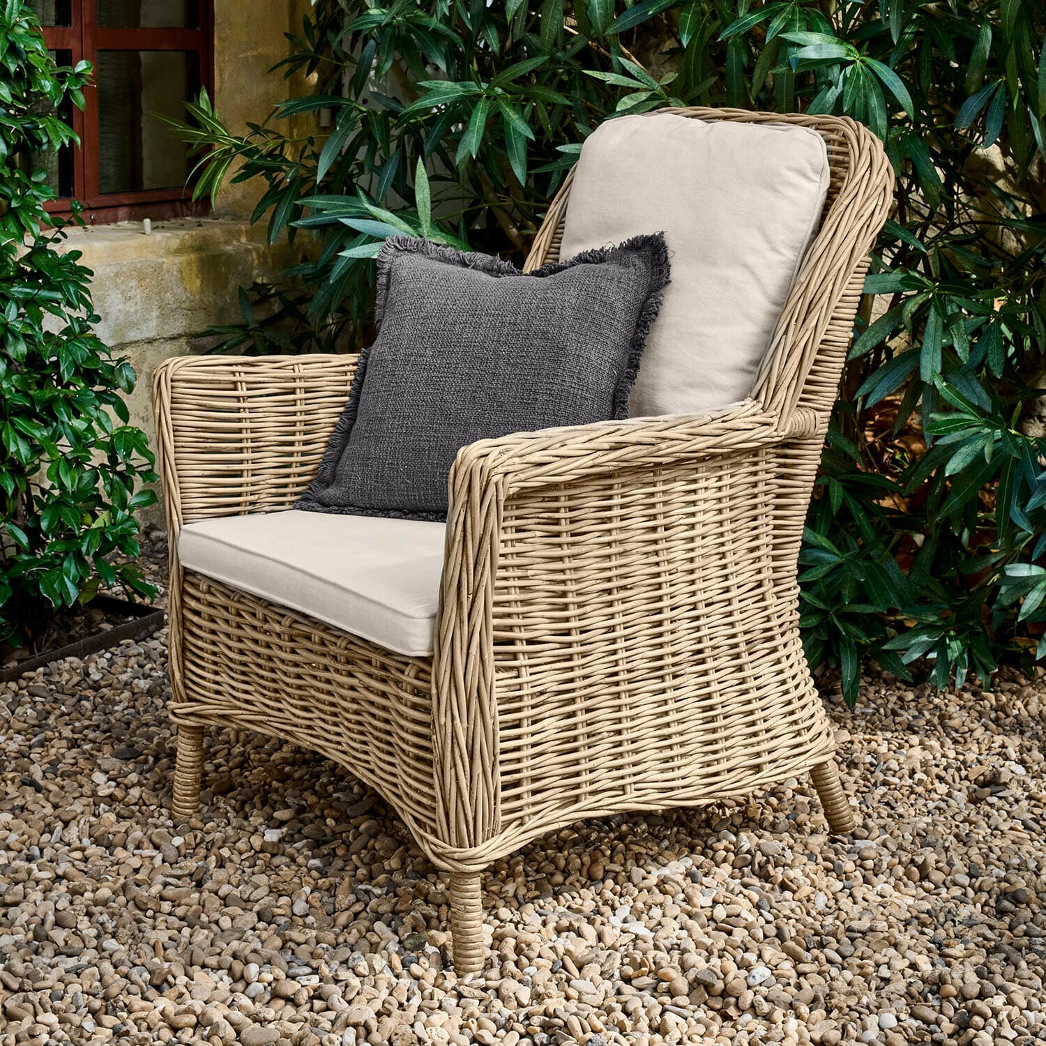 Fauteuil Travenne Rotan fauteuil met crèmekleurige kussens en donkergrijs sierkussen op grindterras