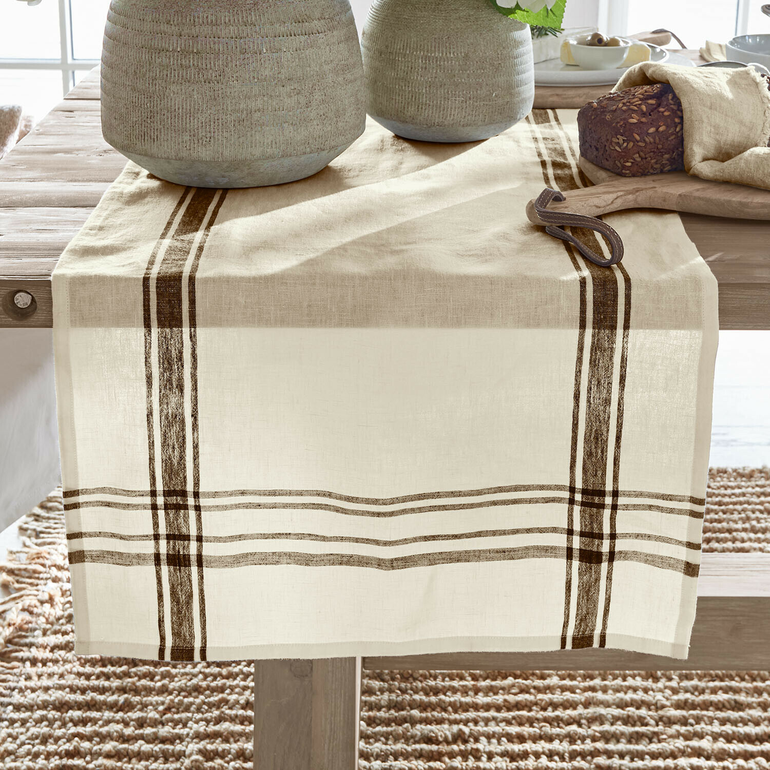 Beige linnen tafelloper met bruine strepen op houten tafel, gedecoreerd met brood en servies