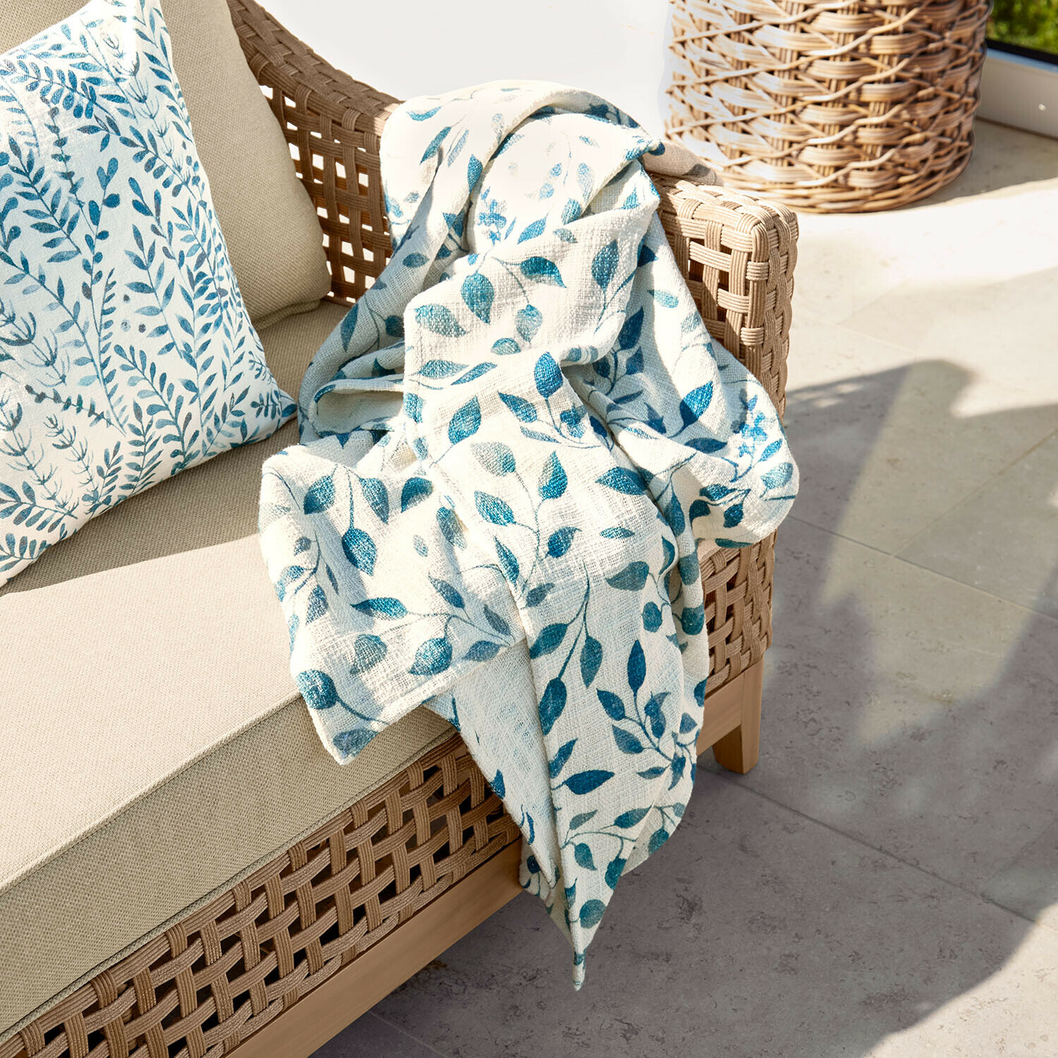 Plaid Ocevara Beige bank met gevlochten rugleuning en crème deken met blauw bladmotief op terras