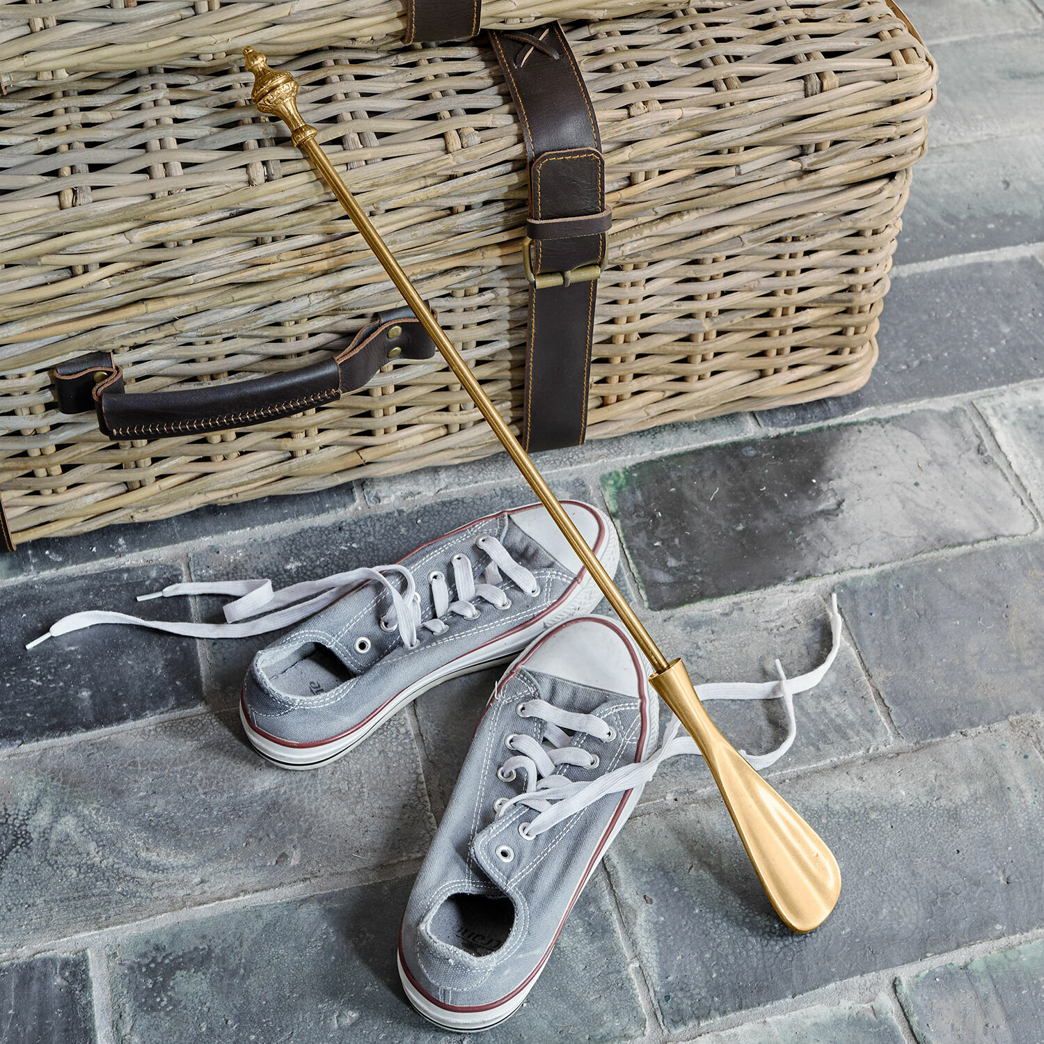 Schoenlepel Orveline Gouden metalen schoenlepel op stenen vloer bij grijze sneakers en een rieten mand