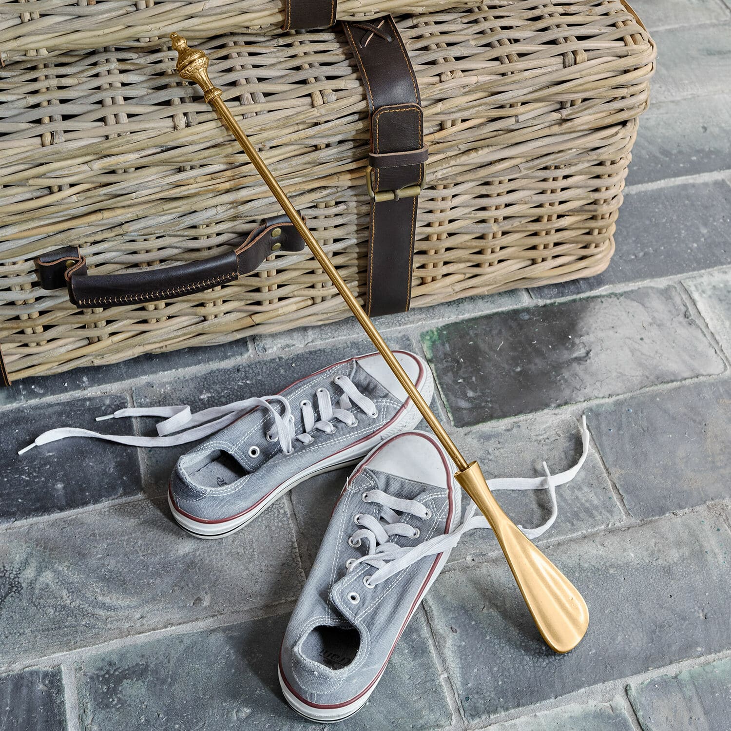 Schoenlepel Orveline Gouden metalen schoenlepel op stenen vloer bij grijze sneakers en een rieten mand