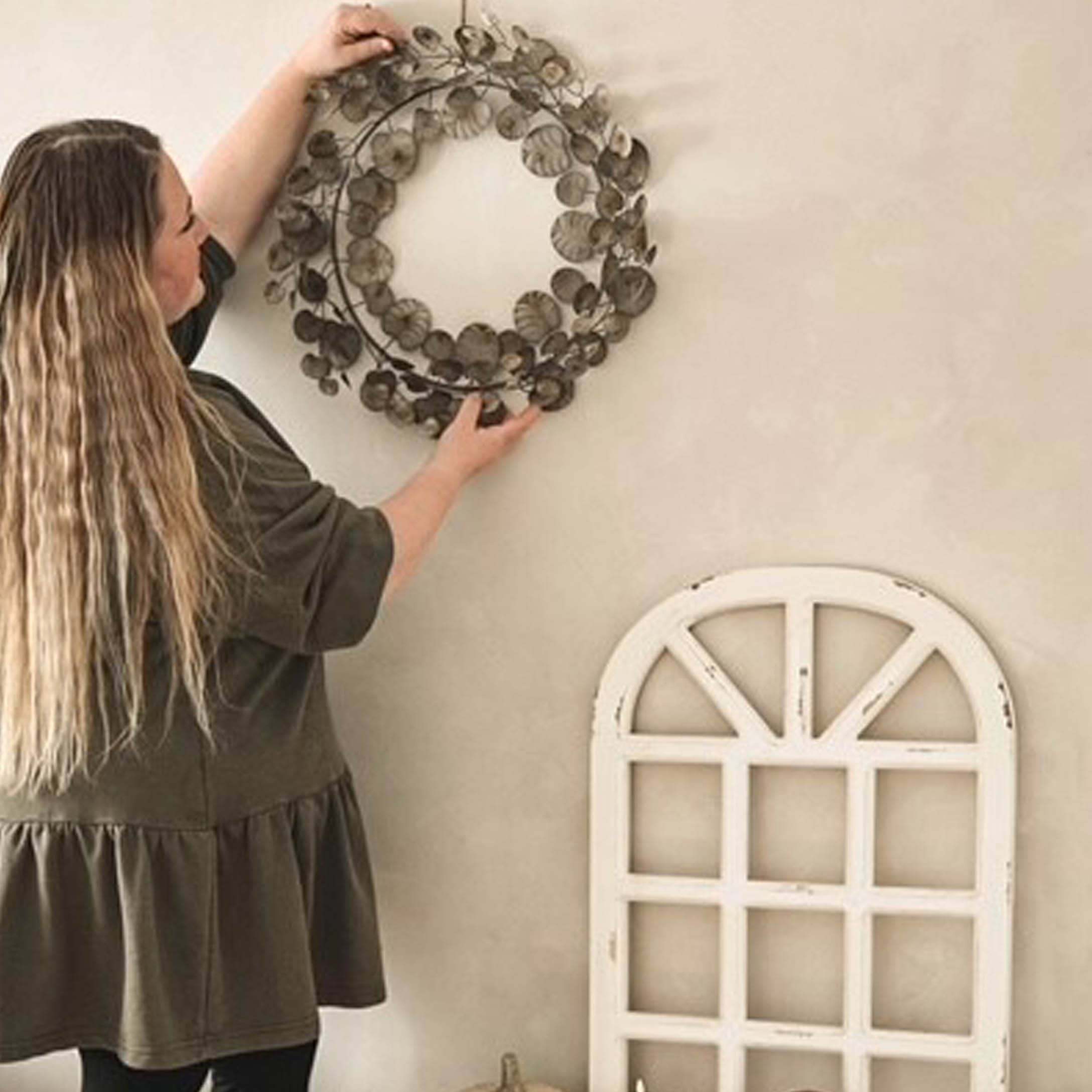 Vrouw hangt bruine metalen decoratieve krans aan lichte muur in rustieke stijl