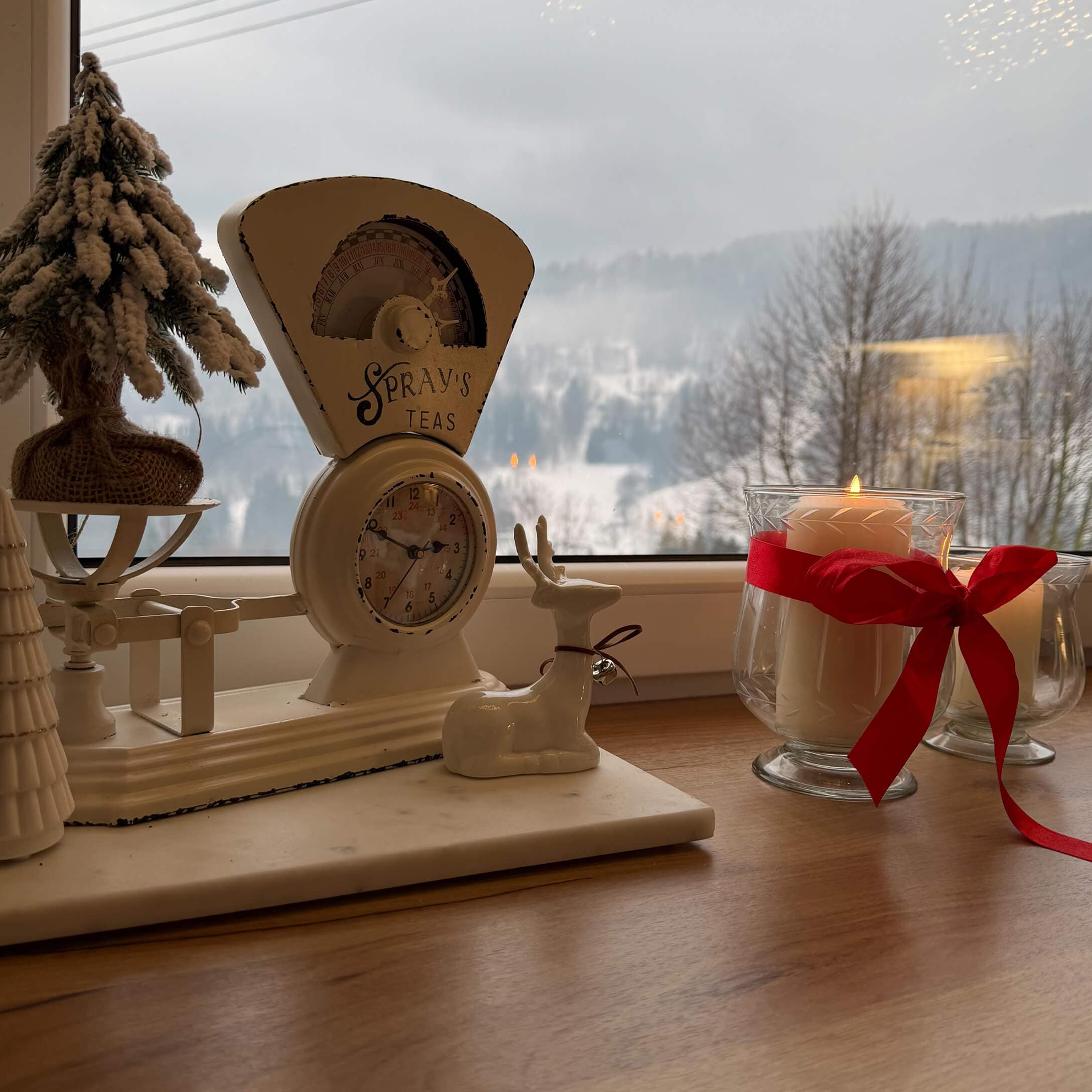 Witte metalen keukenweegschaal met opschrift Spray's Teas, kerstdecoratie en kaars met rode strik op vensterbank