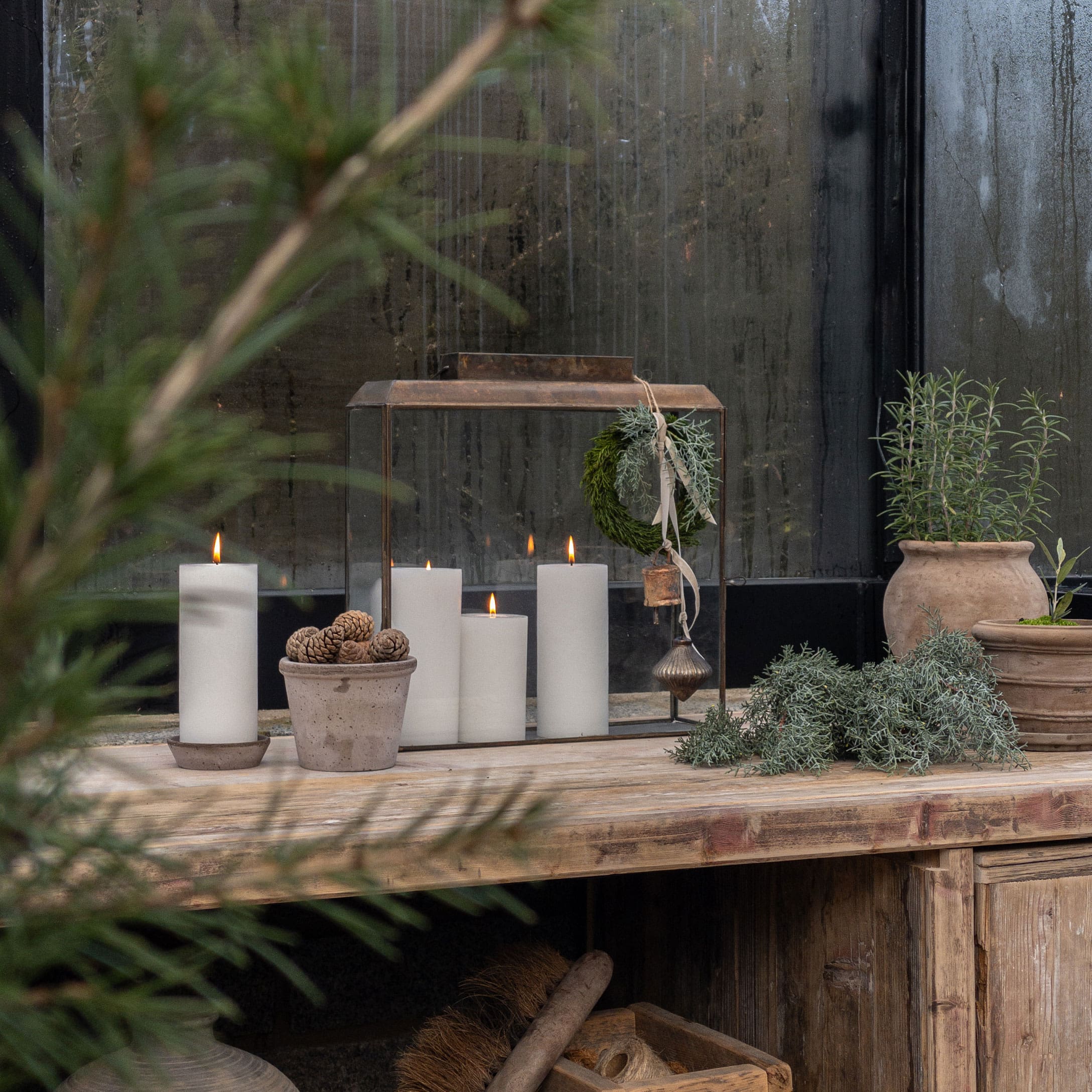 Rustieke houten tafel met witte kaarsen en potten met groene planten voor een beslagen raam