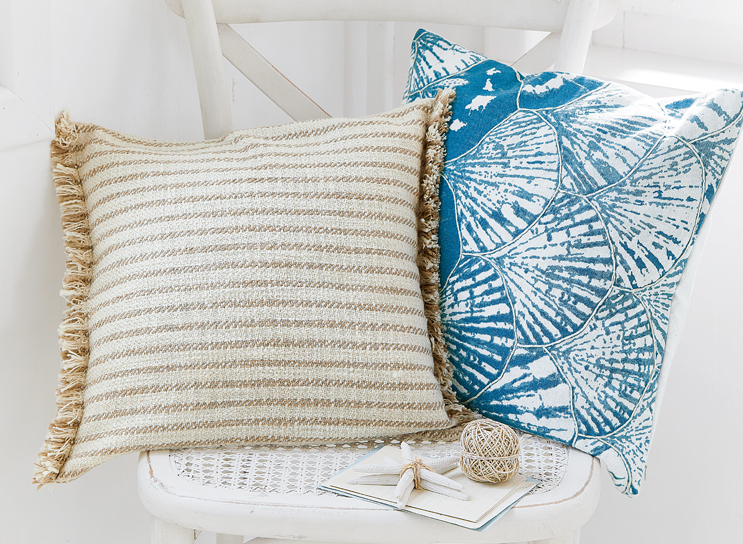 Twee decoratieve kussens op een witte stoel, één beige gestreept met franjes, de ander blauw met schelpenpatroon