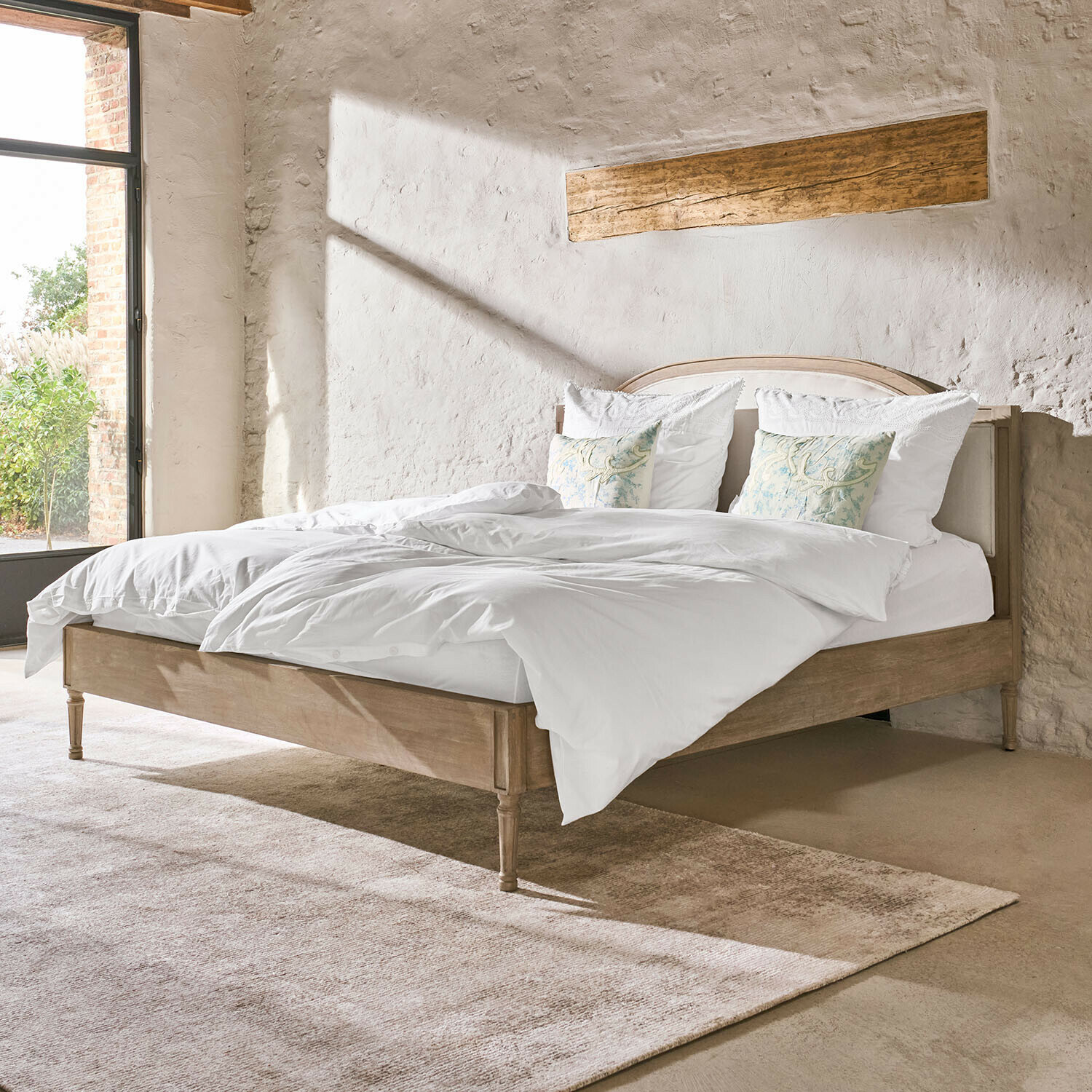 Houten bed in natuurlijke kleur met wit beddengoed en decoratieve kussens in lichte rustieke slaapkamer