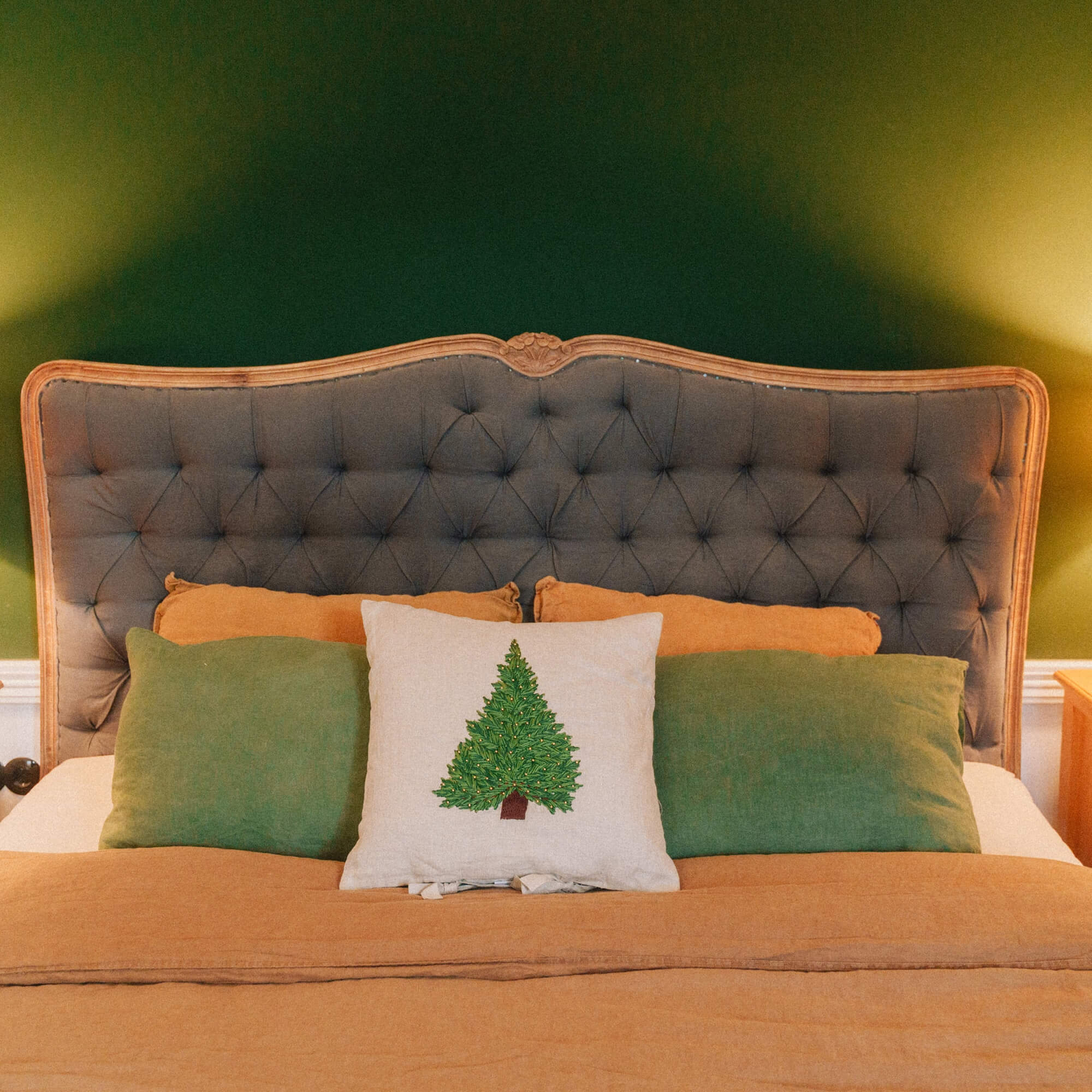 Bed met grijs gestoffeerd hoofdeinde, groene en oranje kussens en kerstkussen met kerstboom