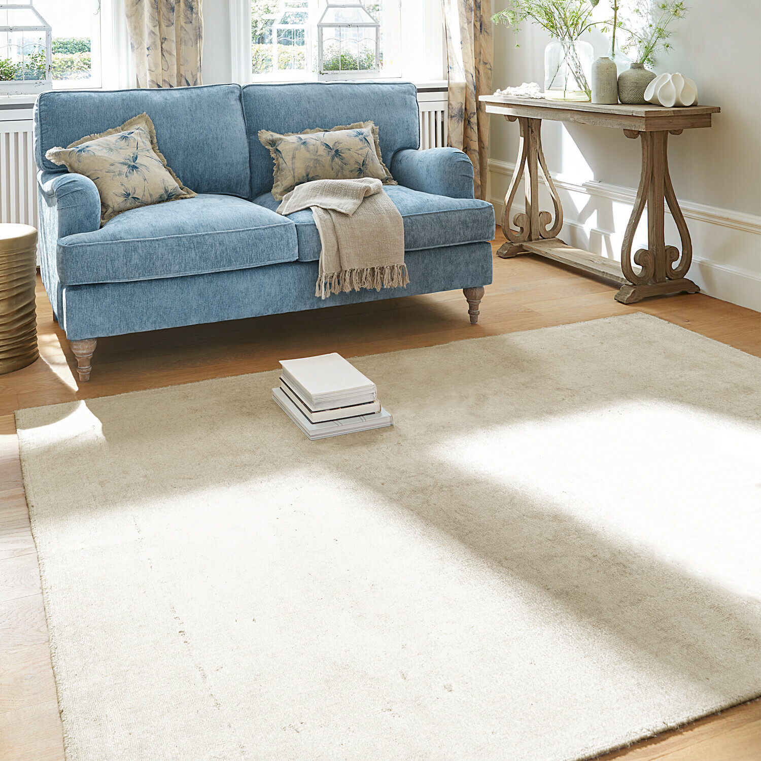 Blauwe stoffen bank met beige deken en kussens, lichte vloerkleed op houten vloer in lichte woonkamer