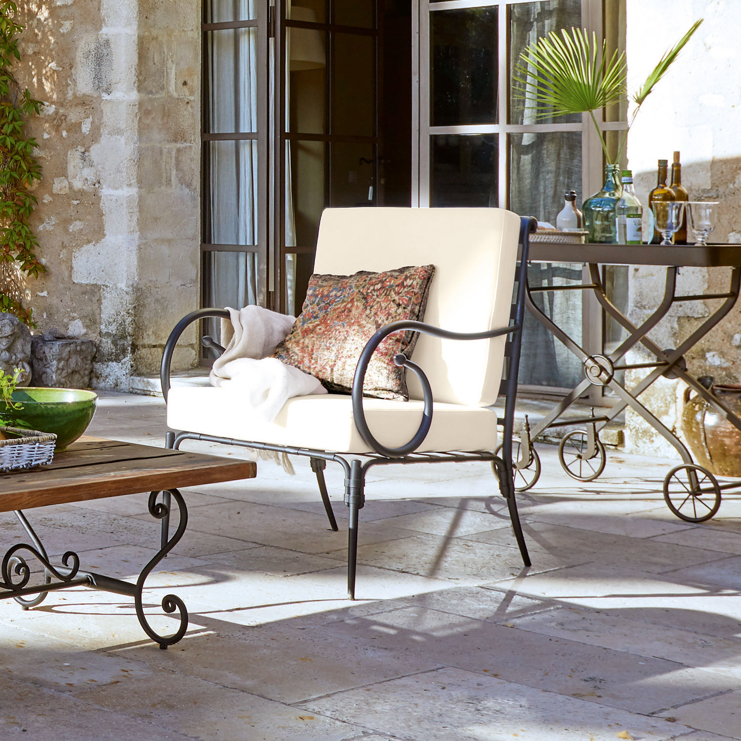 Stoel Neret Metalen tuinbank met crèmekleurige kussens en decoratief kussen op stenen terras