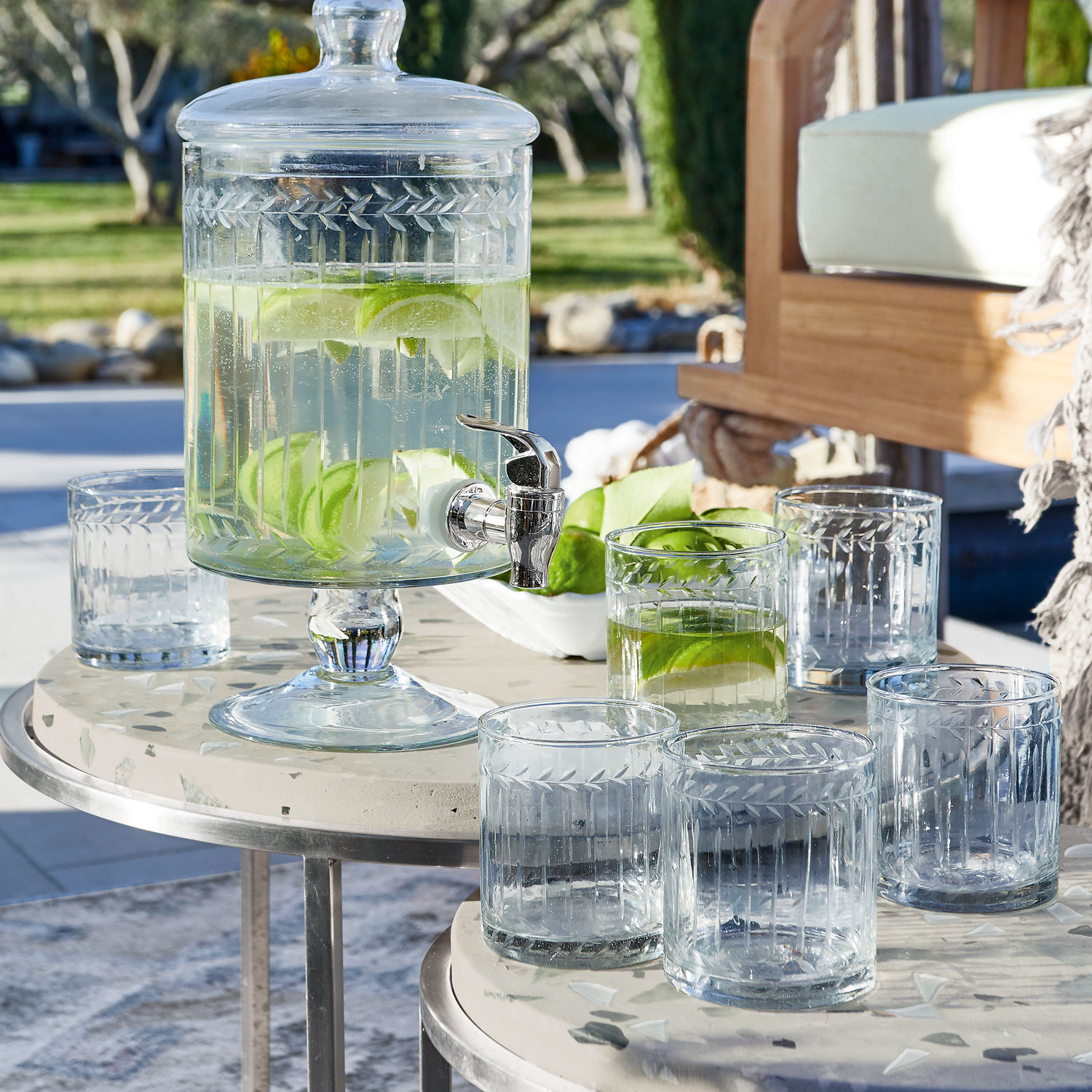 Glazen drankdispenser met deksel en kraan, gevuld met water en limoenpartjes op een terras
