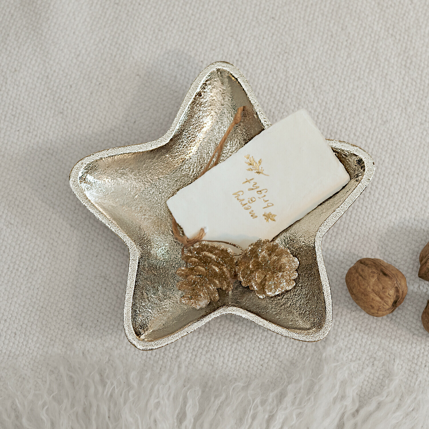 Decoratieve stervormige schaal van zilverkleurig metaal met gouden dennenappels en noten op lichte stof