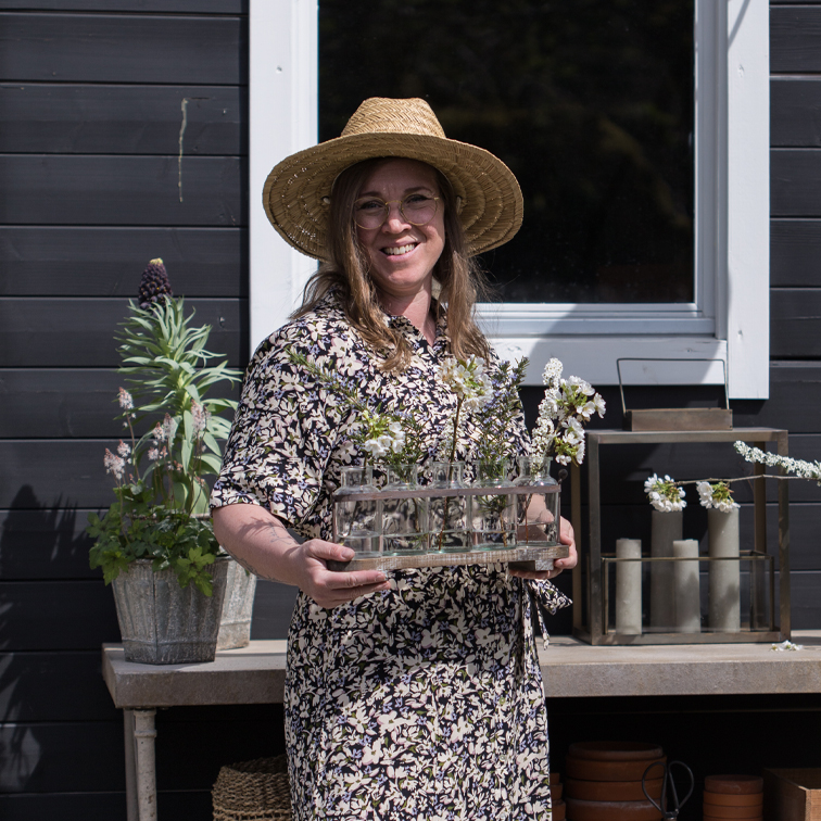 Vrouw met hoed houdt glazen vaas met bloemen voor zwarte muur en raam