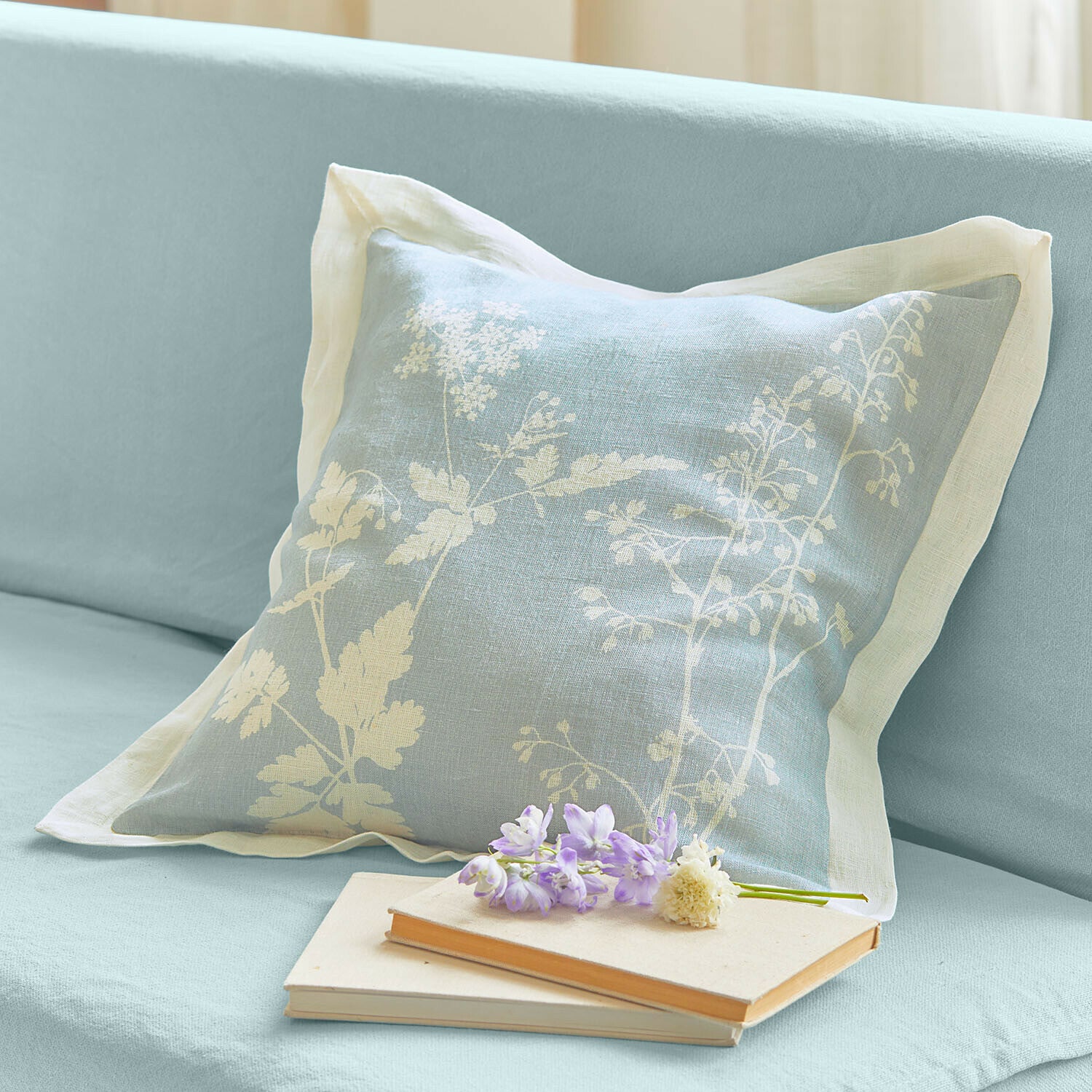 Linnen kussen lichtblauw met wit bloemenpatroon op bank, met boeken en bloemen ernaast
