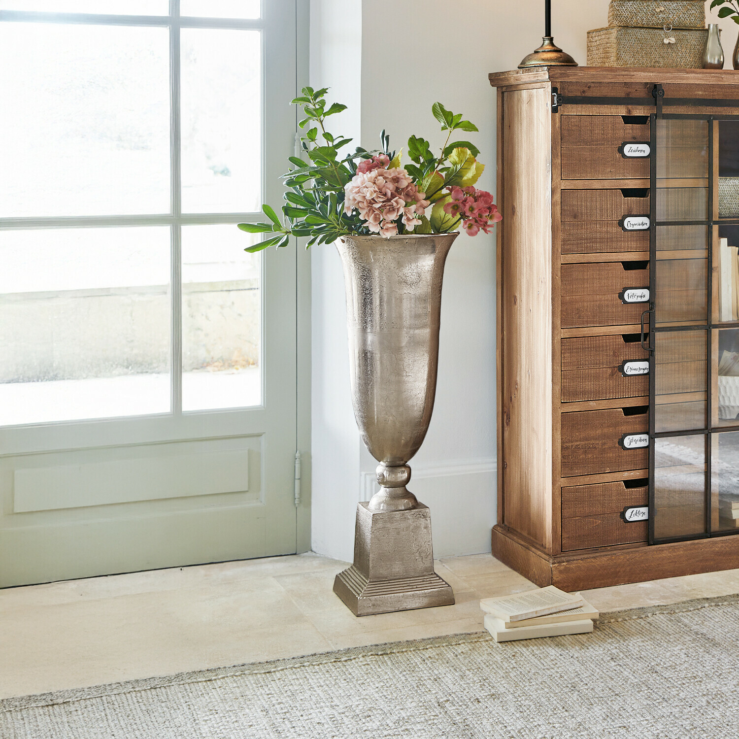 Vaas Belfort Hoge zilverkleurige metalen vaas met bloemen naast een houten kast in lichte kamer
