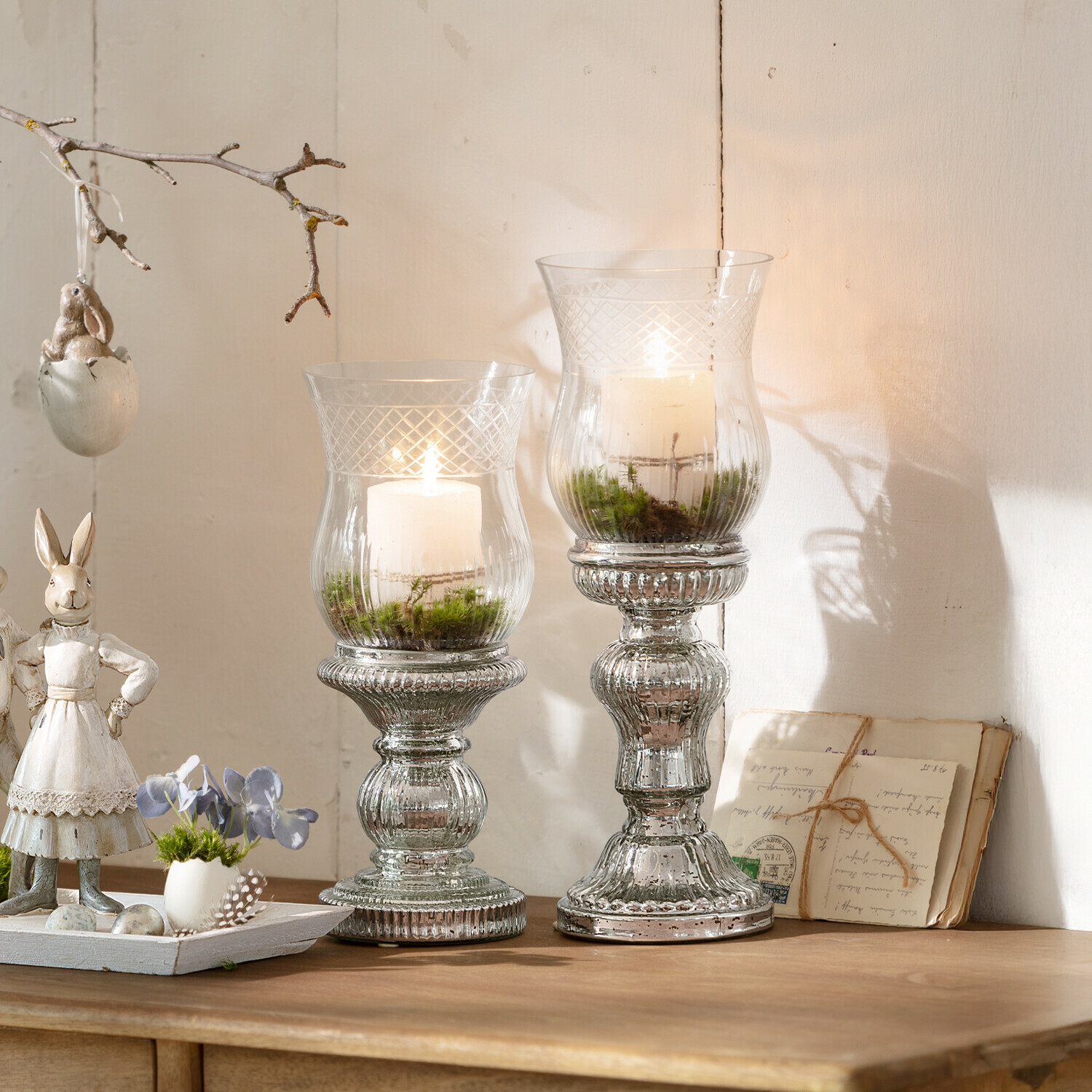 Twee glazen kandelaren met witte kaarsen en groen mos op houten tafel, lentebloemstuk