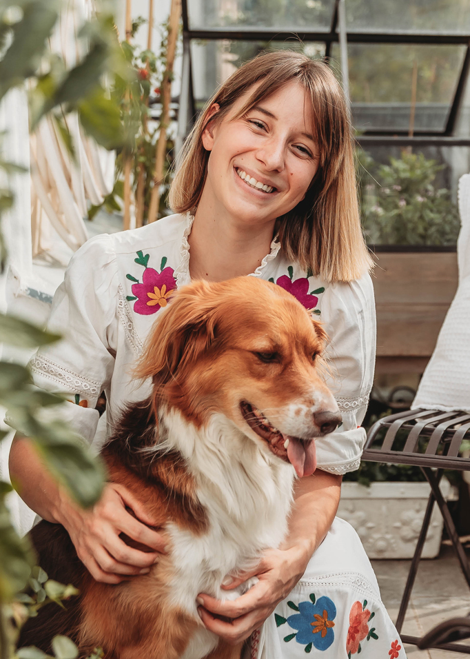 Vrouw in witte jurk met kleurrijk borduurwerk houdt bruin-witte hond vast in lichte kamer met planten