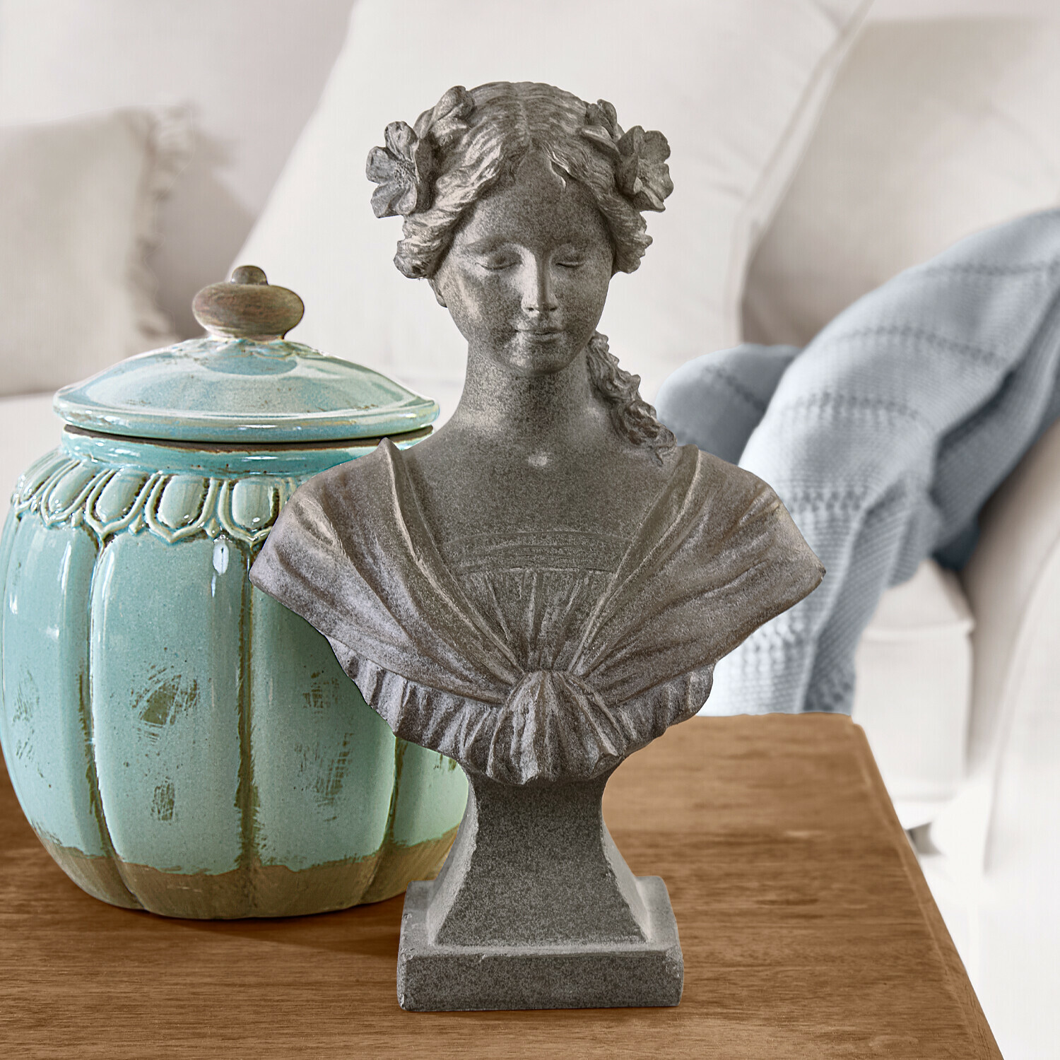 Grijs stenen vrouwenbuste met bloemen in het haar op houten tafel naast turquoise keramische pot