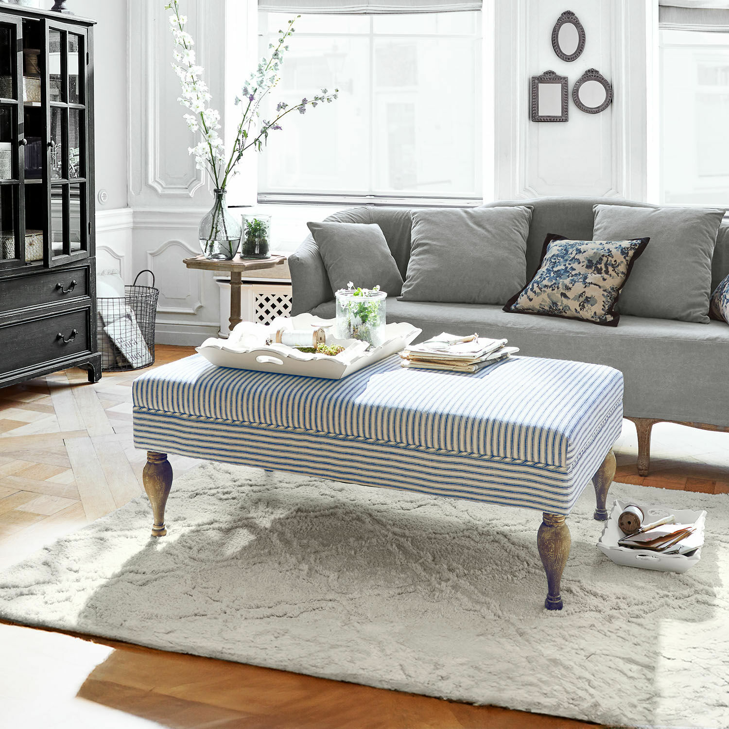 Gestreepte poef met houten poten op lichte vloerkleed in moderne woonkamer met grijze bank