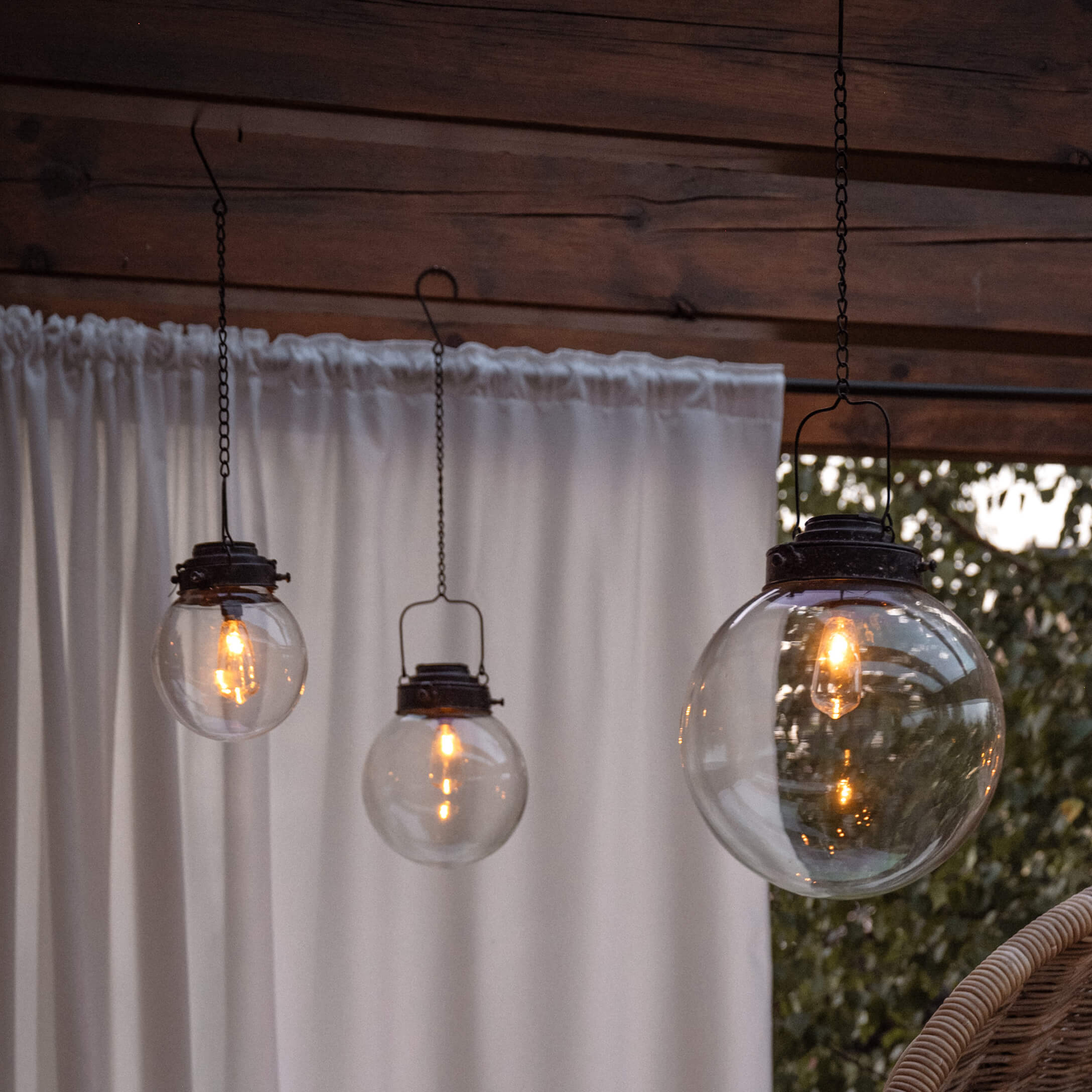 Drie hanglampen van helder glas met zwarte metalen onderdelen op terras met wit gordijn