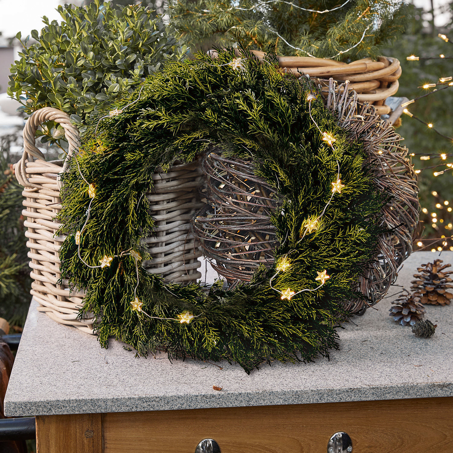 Krans Évres Groene kerstkrans van takken met stervormige lichtjes op stenen blad naast rieten manden