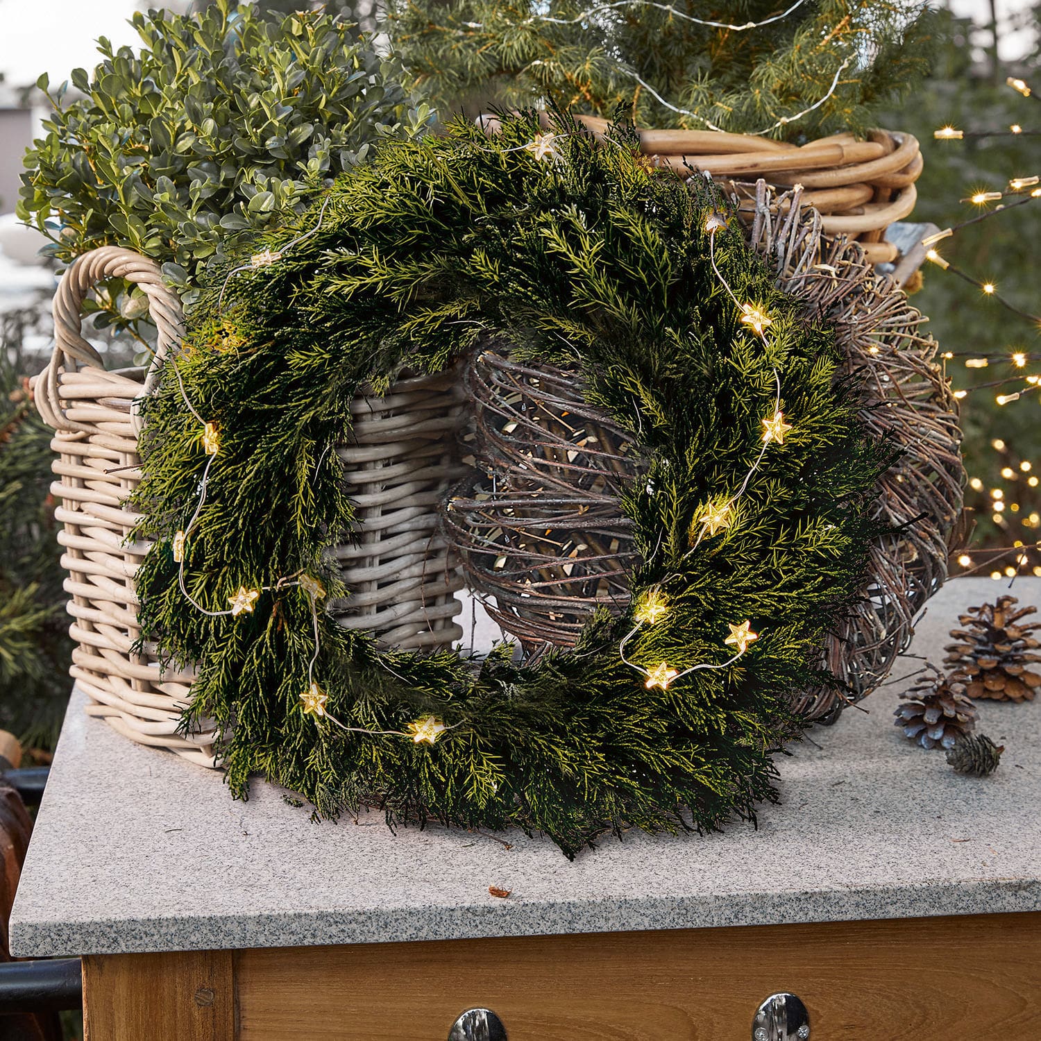 Krans Évres Groene kerstkrans van takken met stervormige lichtjes op stenen blad naast rieten manden