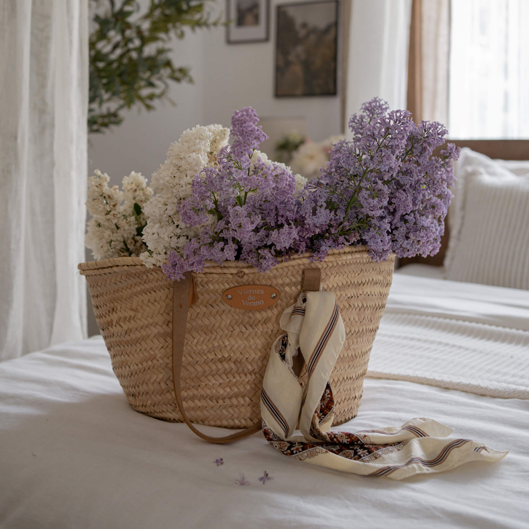Gevlochten rieten mand met witte en paarse bloemen en sjaal op wit bed in lichte kamer