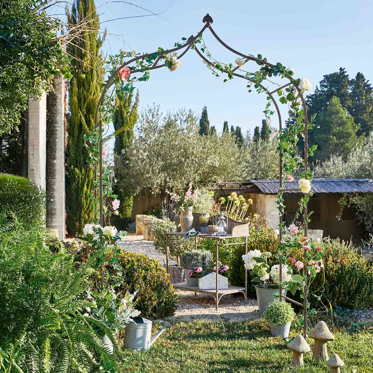 Metalen tuinpergola met groene bladeren en roze bloemen in zonnige tuin