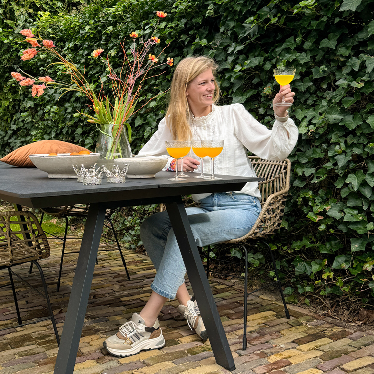 Vrouw in witte blouse en spijkerbroek zit buiten aan zwarte tafel, houdt glas met oranje drankje vast, vaas met bloemen ernaast