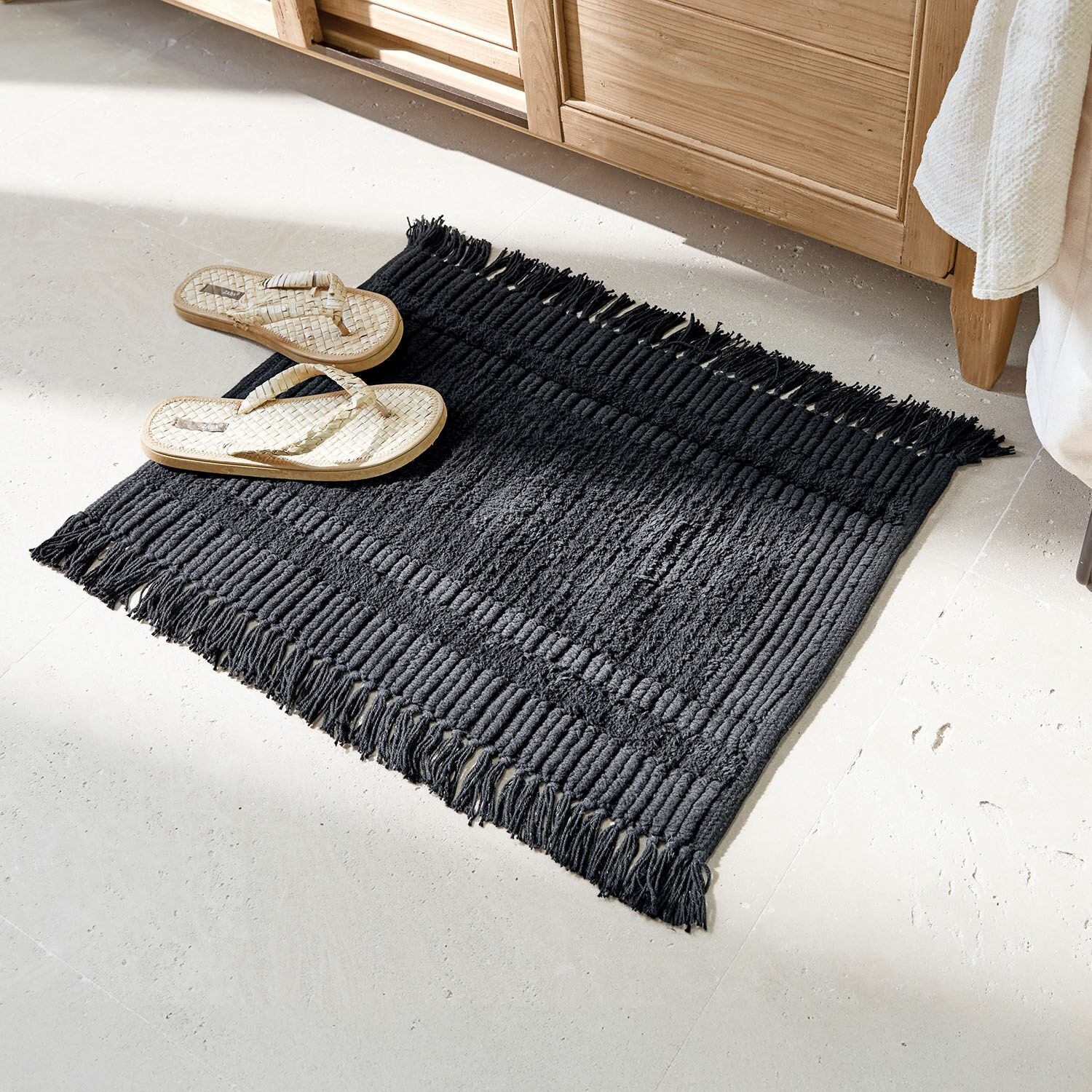 Badmat Nexilo Zwart katoenen vloerkleed met franjes op lichte vloer, naast lichte gevlochten slippers