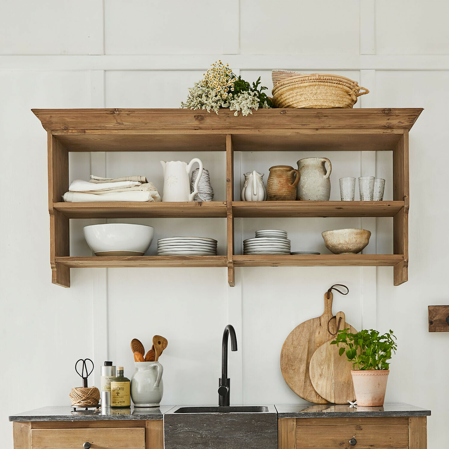 Houten keukenplank met keramisch servies, borden en rieten mand aan witte muur in landelijke stijl