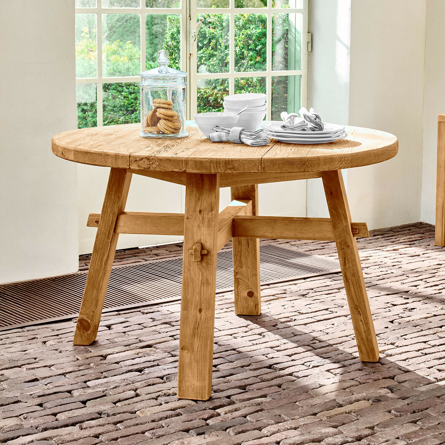 Tafel Crestwooden Ronde tafel van natuurlijk grenenhout op een bakstenen vloer bij een raam met veel licht